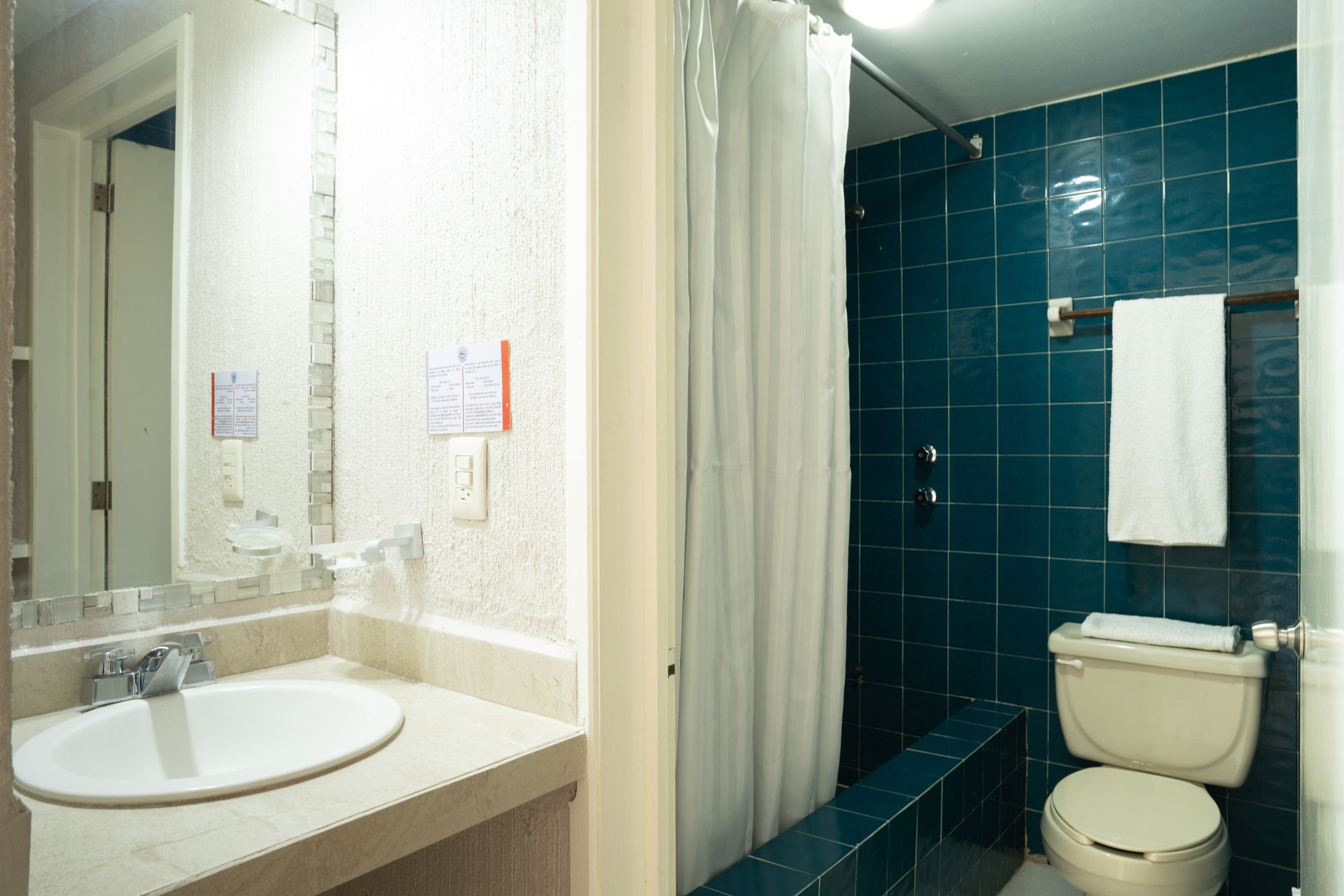Baño con lavabo e inodoro blancos, ducha con azulejos azules y una toalla blanca colgada.