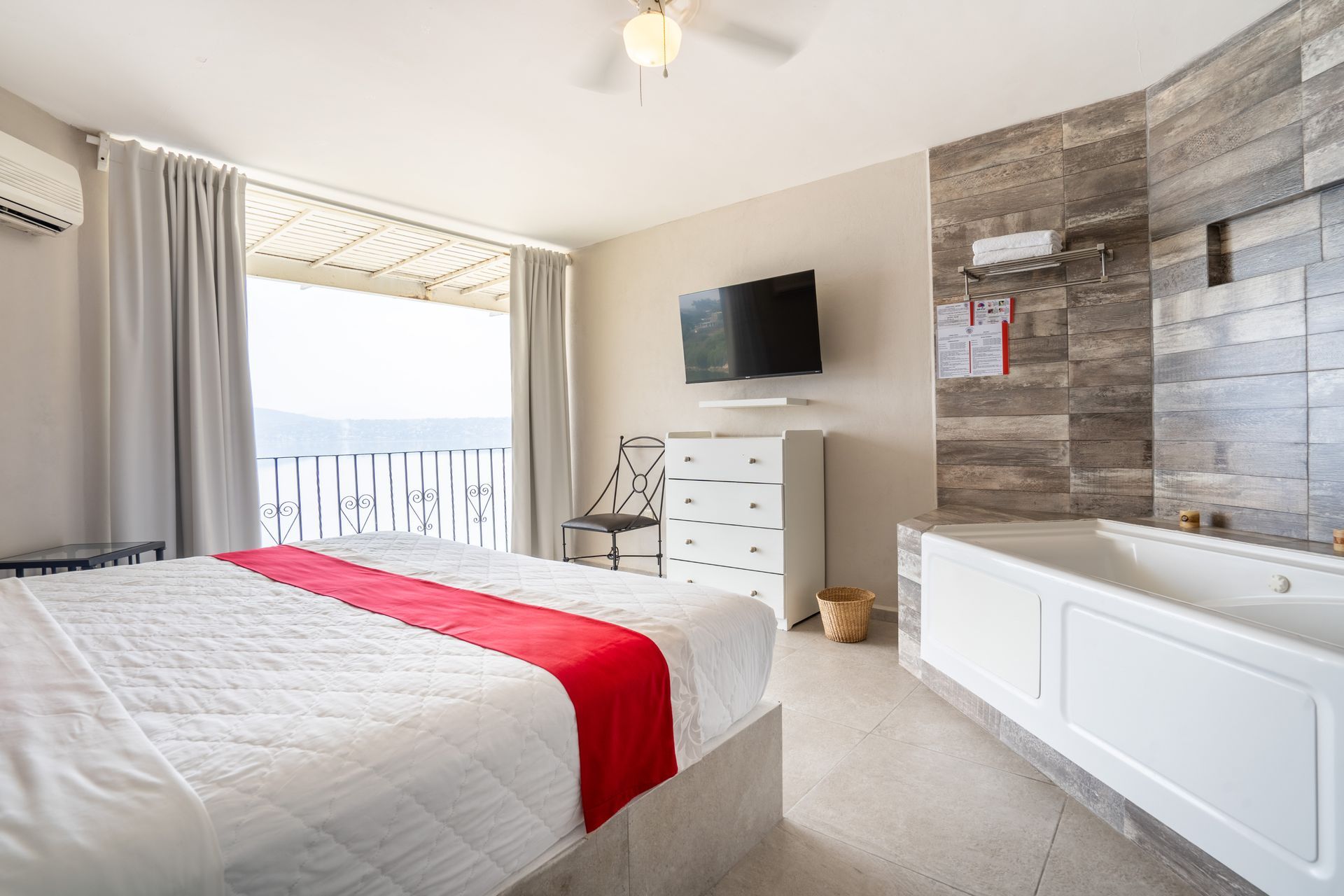 Dormitorio con cama king size, alfombra roja, TV, jacuzzi y balcón con vista al agua.