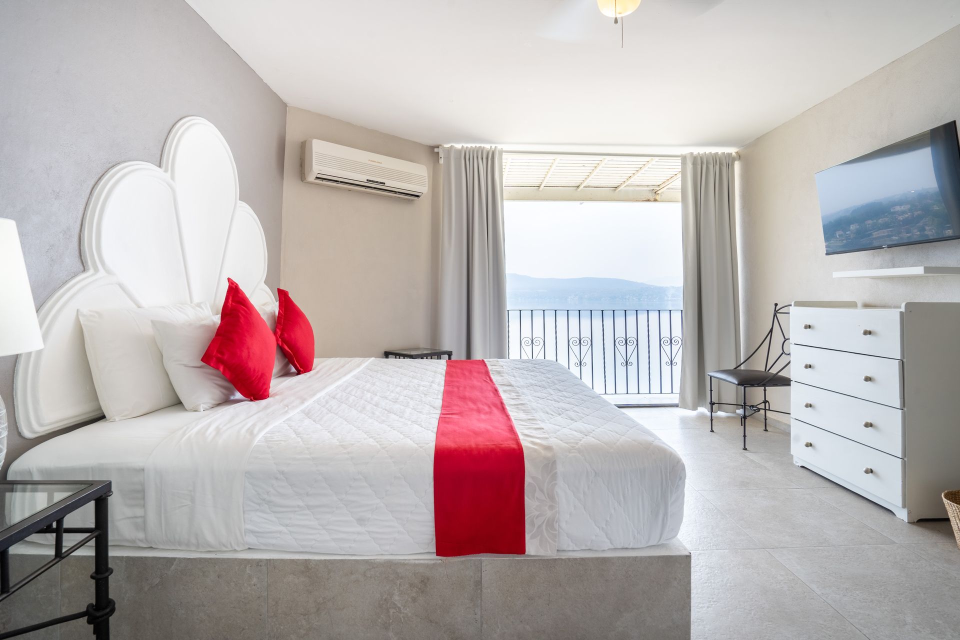Dormitorio con cama king-size, almohadas rojas, balcón, tocador blanco y vista a las montañas y al agua.