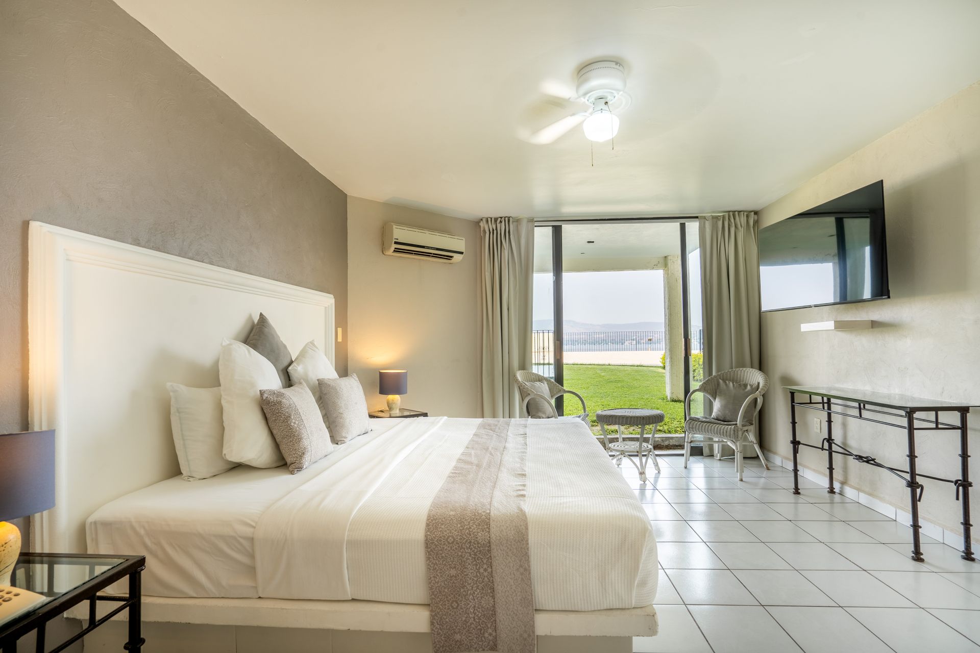 Una luminosa habitación de hotel con cama King, balcón y TV, con vista a una zona verde y al océano.