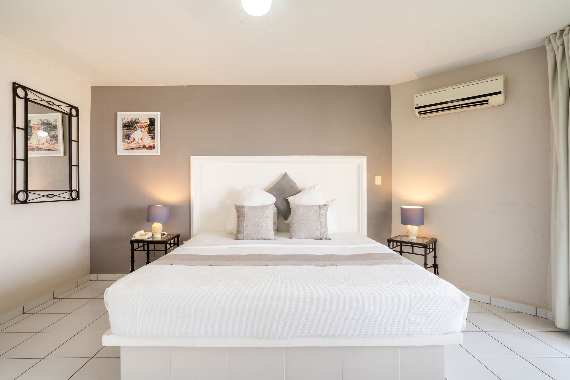 Dormitorio con cama blanca, cabecero gris, lámparas, espejo y aire acondicionado.