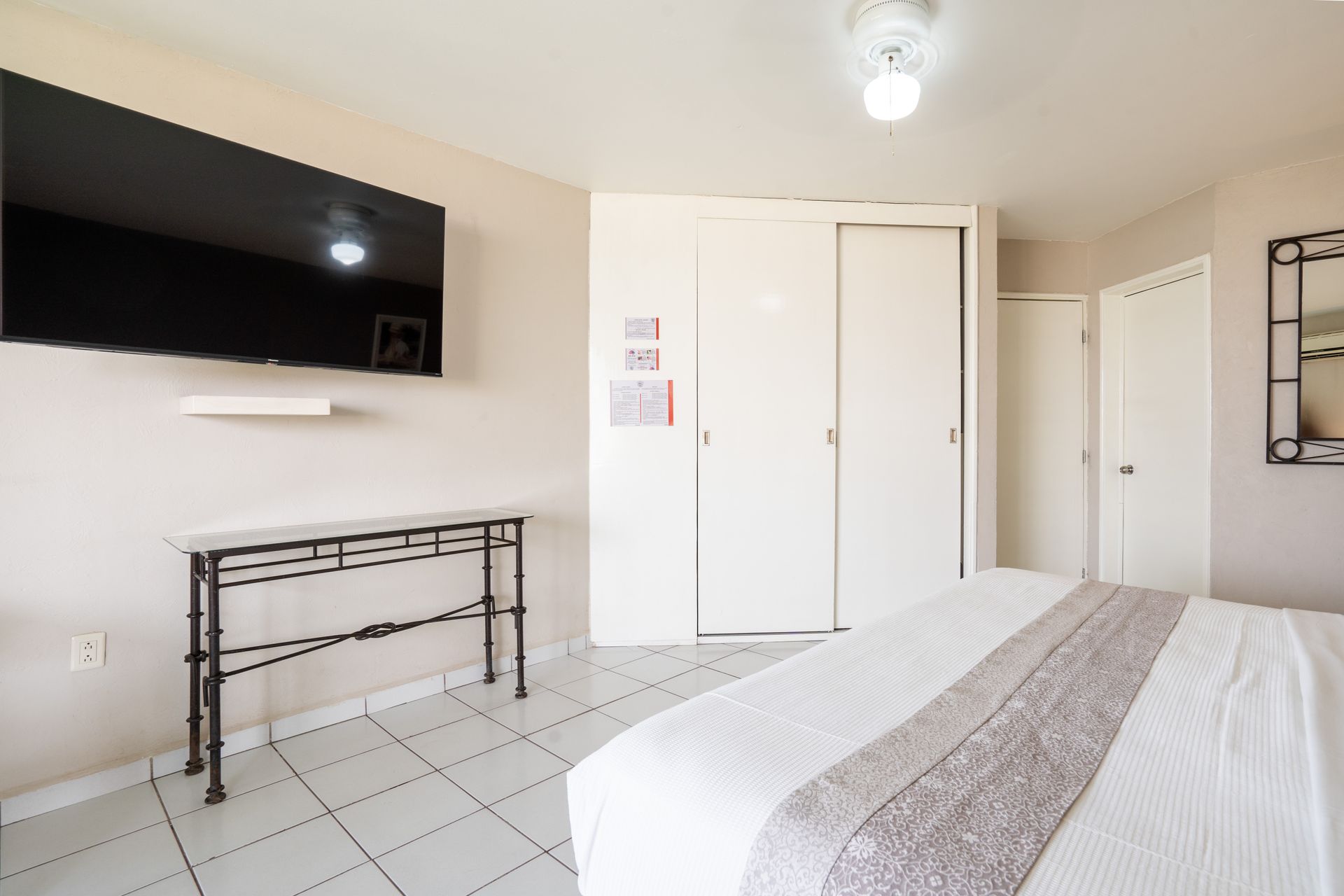 Dormitorio con TV grande, puertas de armario blancas y una cama con alfombra gris.