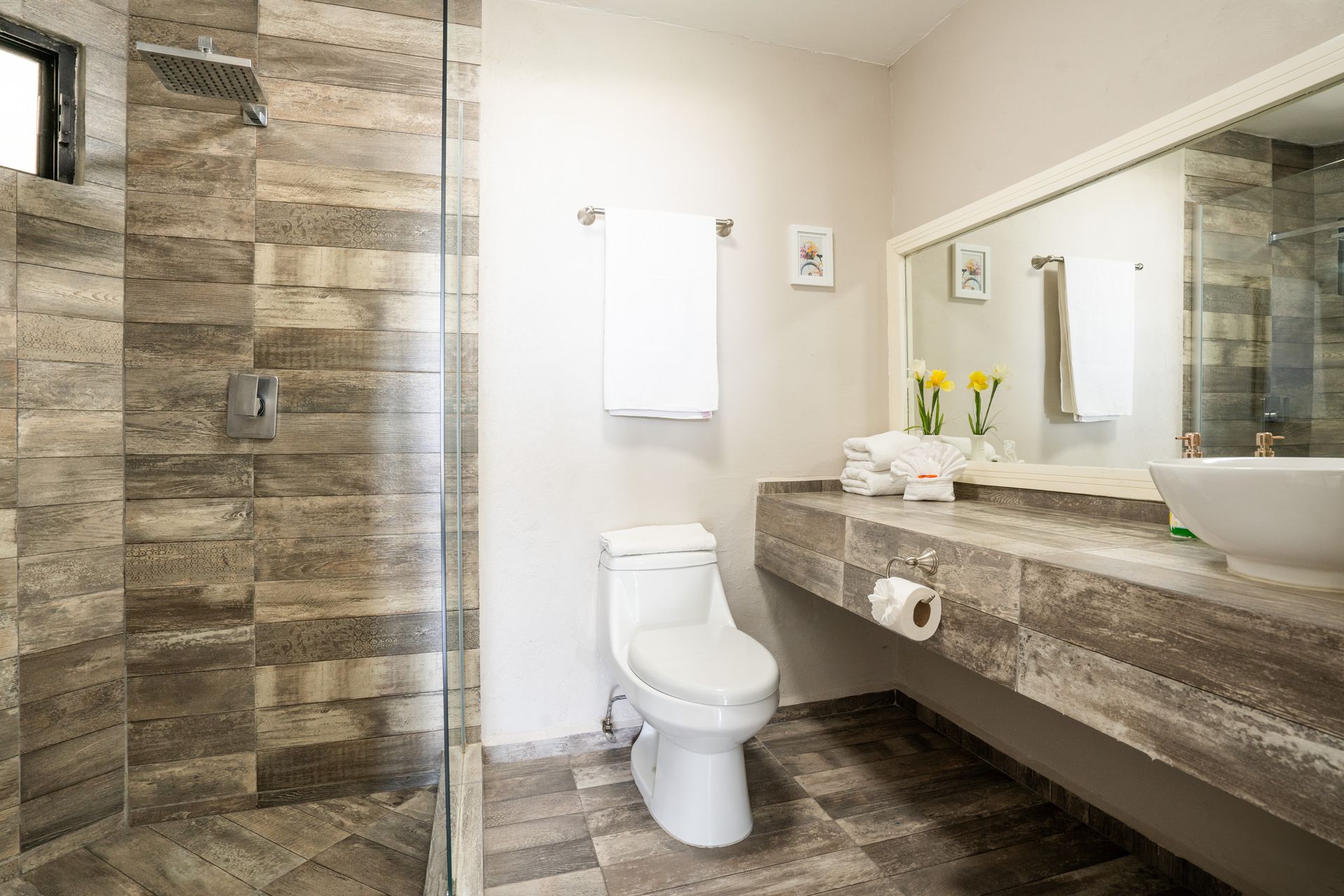 Baño con ducha de azulejos grises, inodoro y lavabo blancos. Un espejo cuelga sobre la encimera y un toallero.