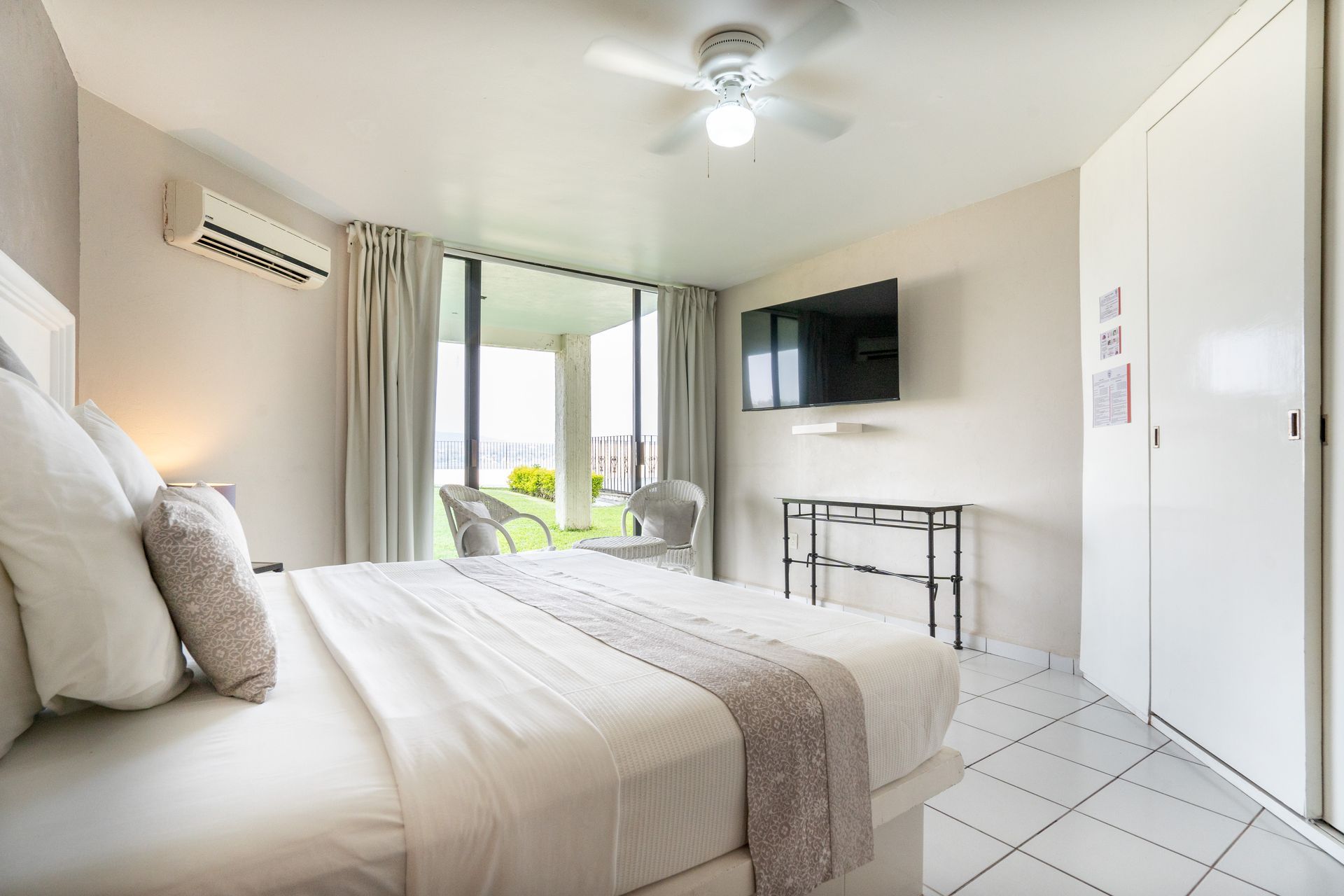 Dormitorio con cama king-size, TV y acceso al patio; tonos neutros con detalles en blanco y gris.