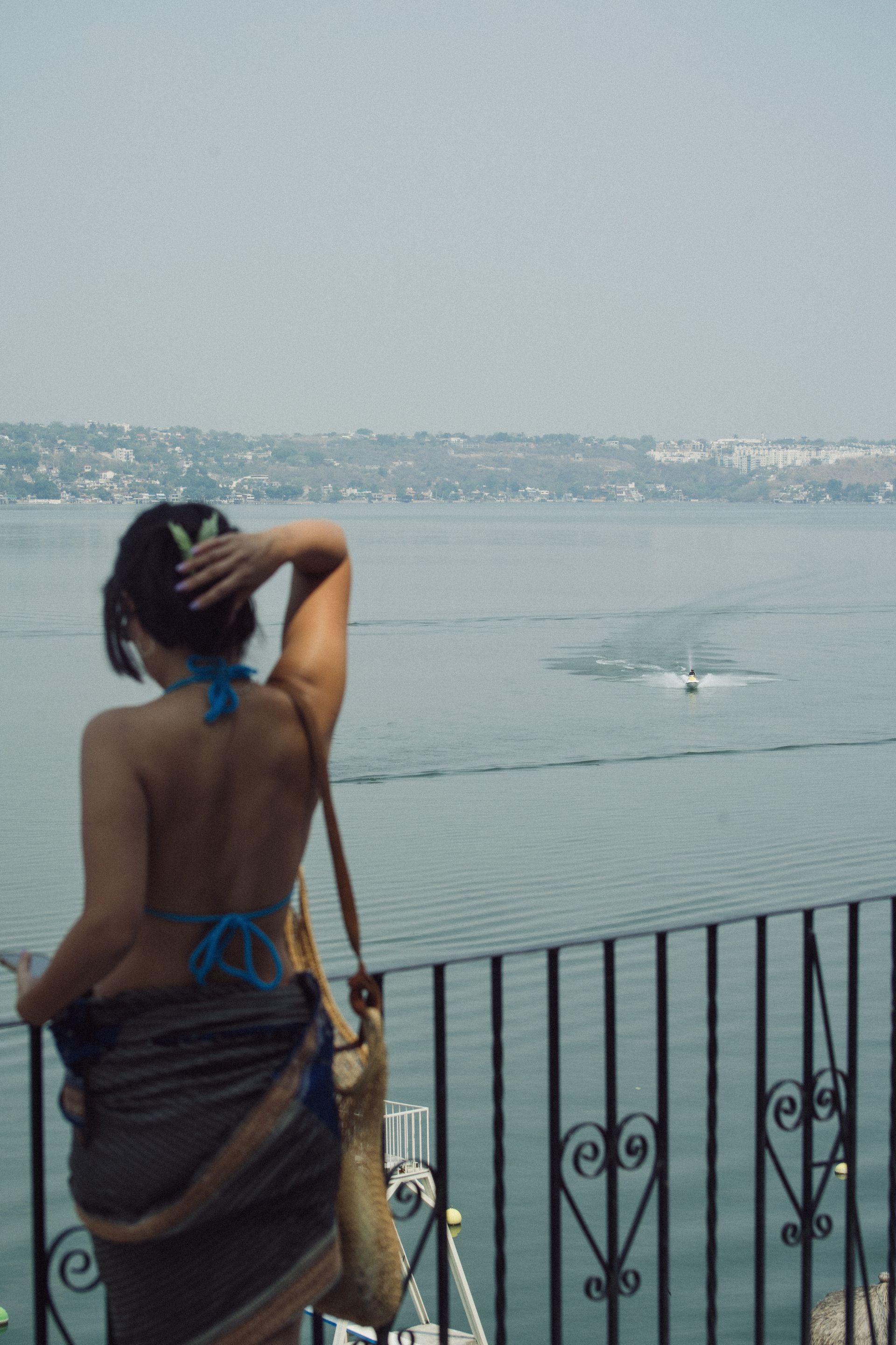 Mujer con la parte superior de un bikini azul mirando hacia un cuerpo de agua, sujetándose el cabello.