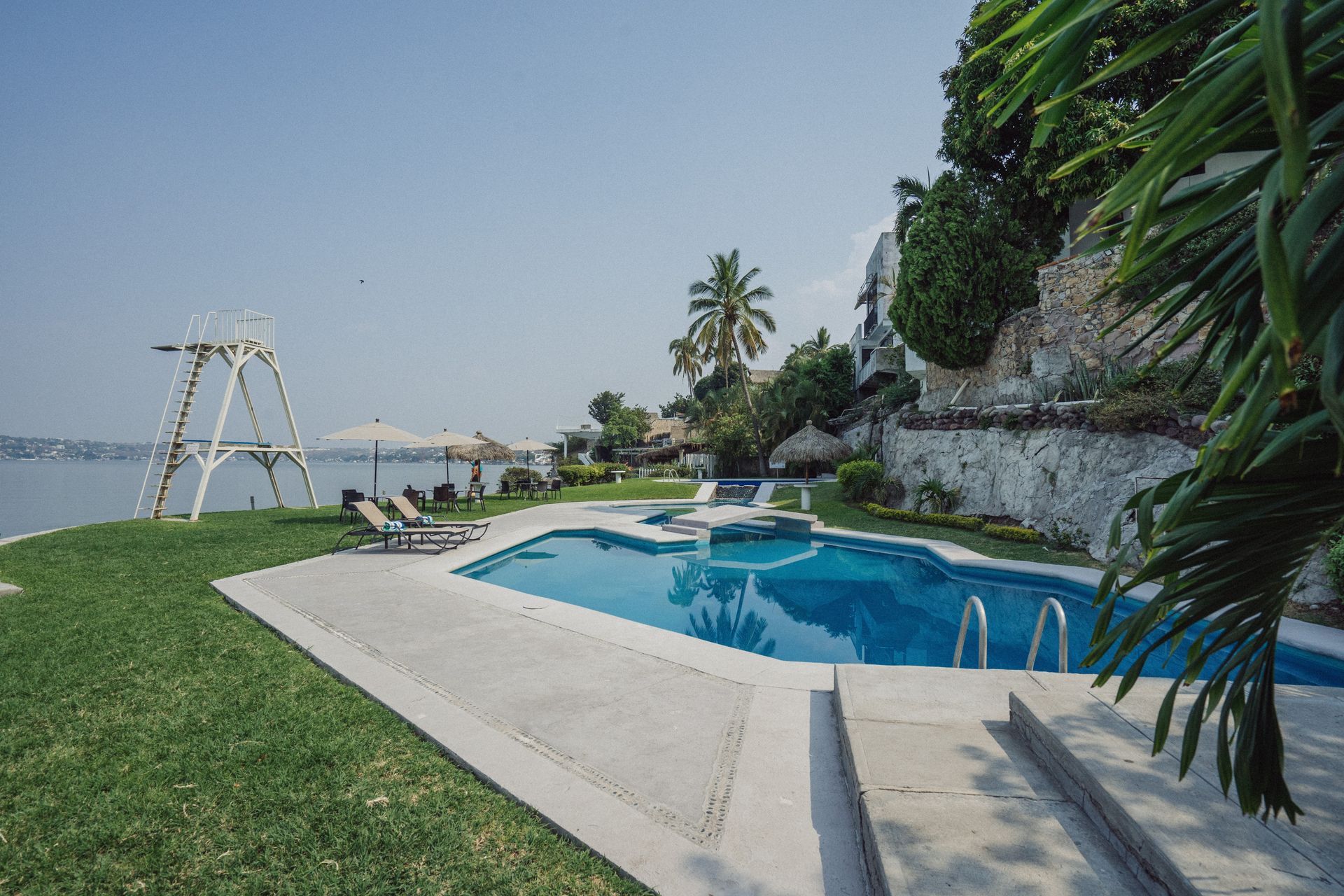Una piscina frente al mar con trampolín, rodeada de césped y palmeras, con un cielo azul brillante.