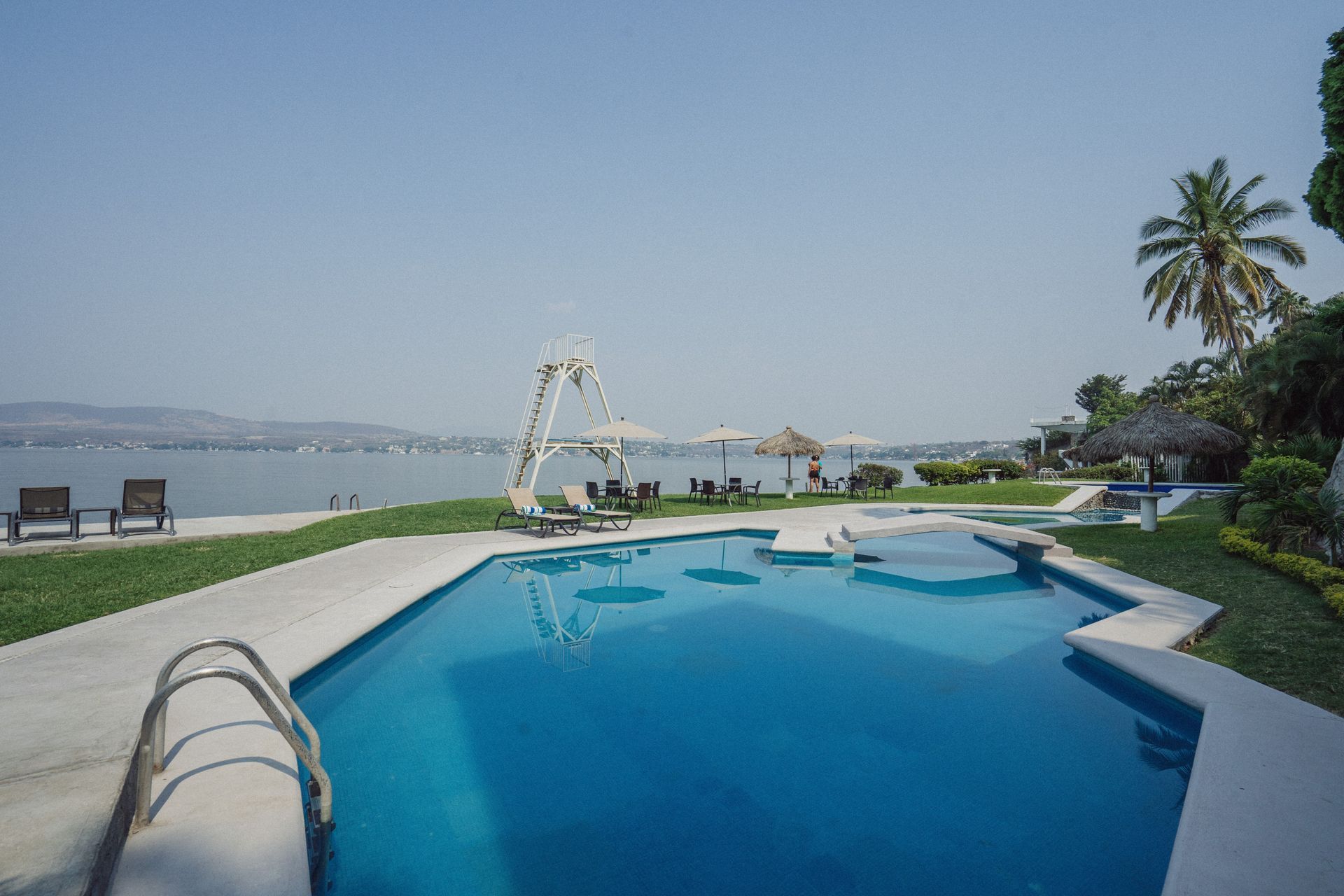 Piscina con vistas a un lago. Agua azul, césped verde y cielo despejado.