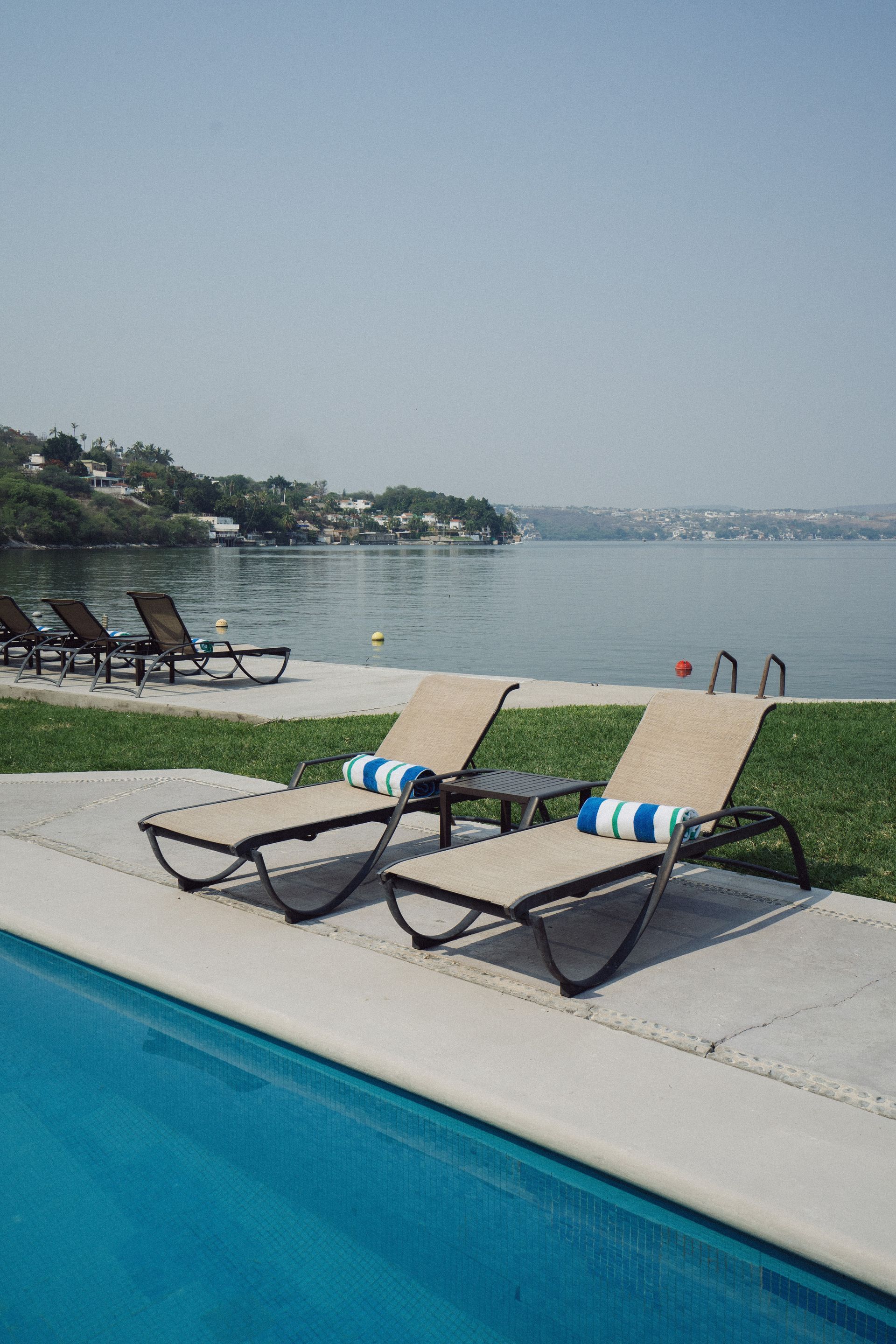 Tumbonas junto a una piscina con vistas al lago. Día soleado con cielo despejado y edificios al fondo.
