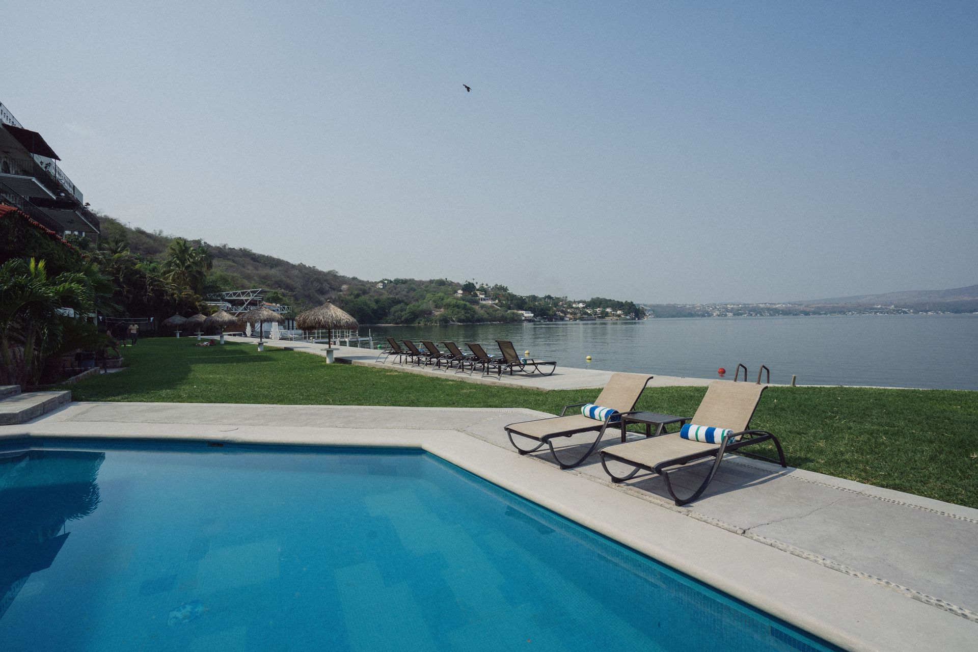 Vista de una piscina, un lago y montañas con tumbonas en un día soleado.