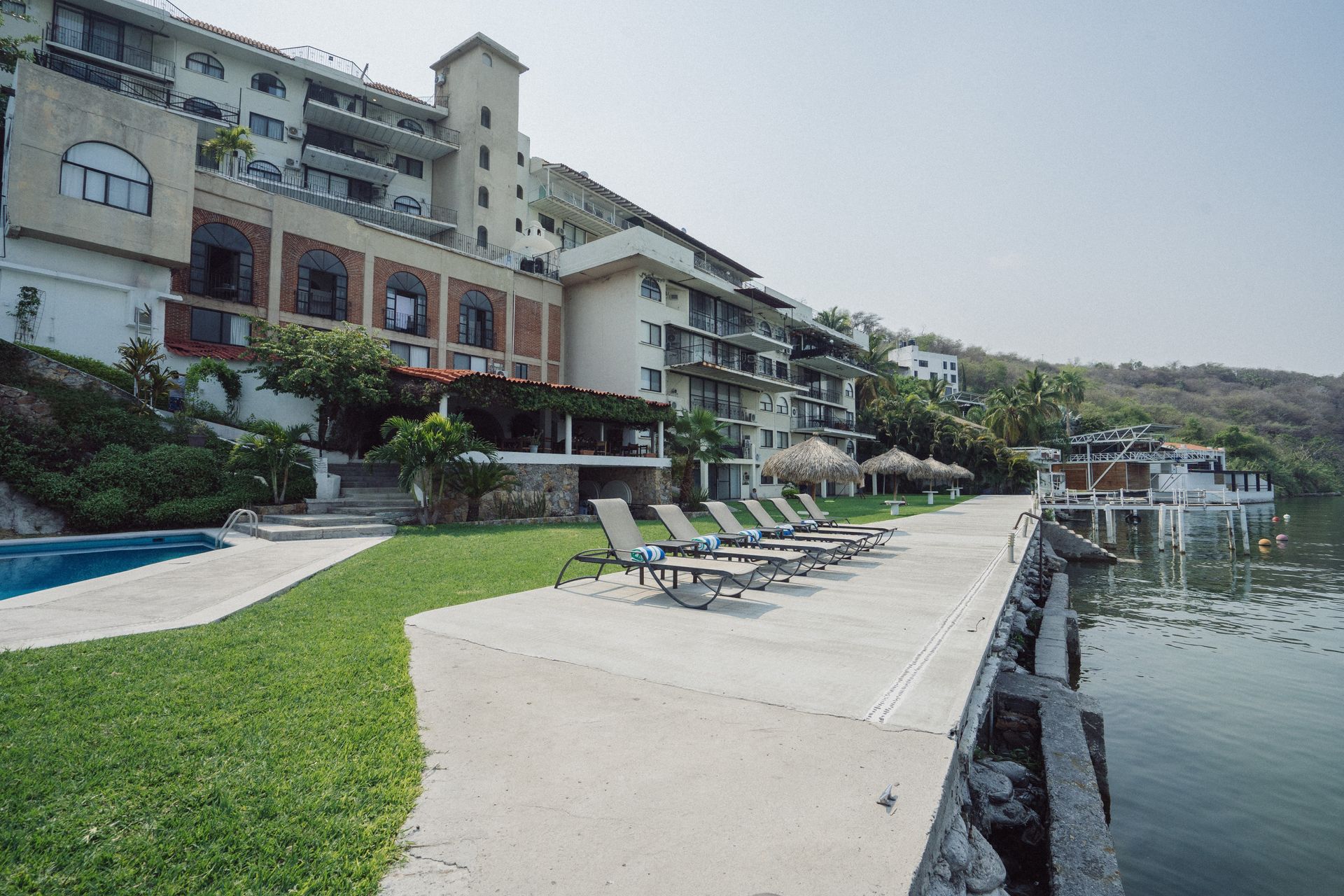 Hotel con piscina y muelle con vista a un lago, reposeras en el muelle y una montaña al fondo.