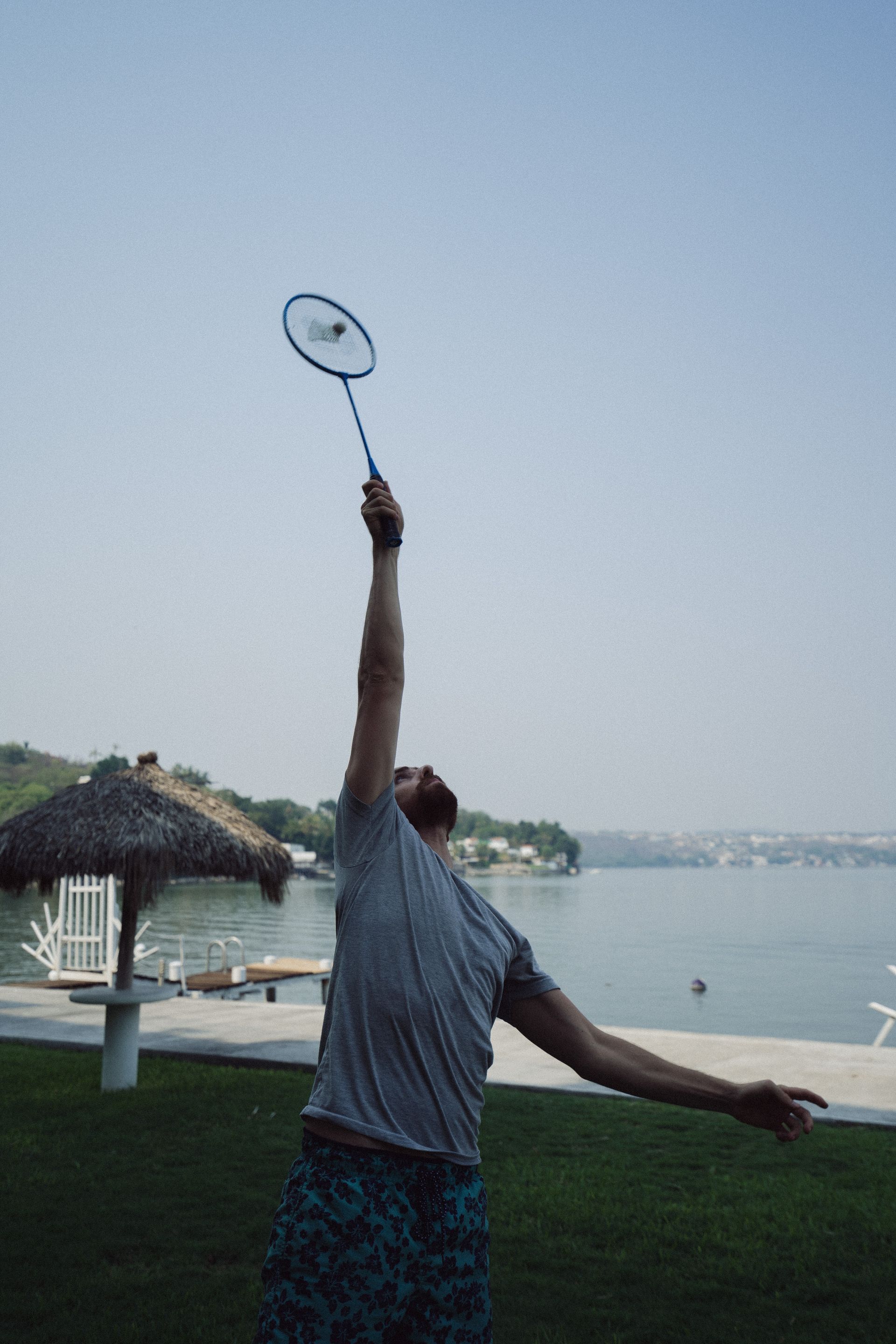 Un hombre con camisa gris balancea una raqueta de bádminton, apuntando hacia el cielo cerca del agua.