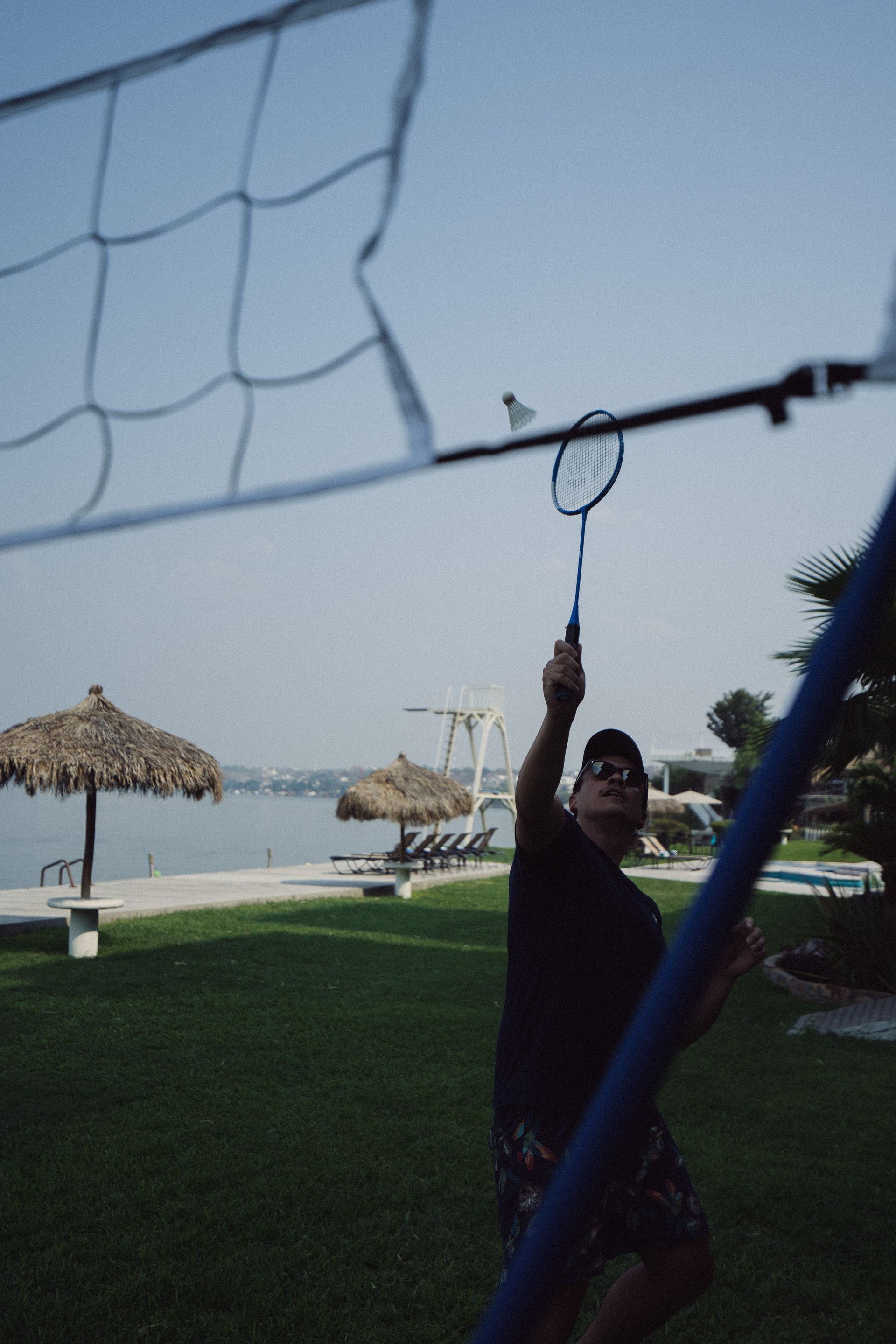 Hombre jugando bádminton cerca de un lago, golpeando el volante por encima de una red, bajo un cielo soleado.