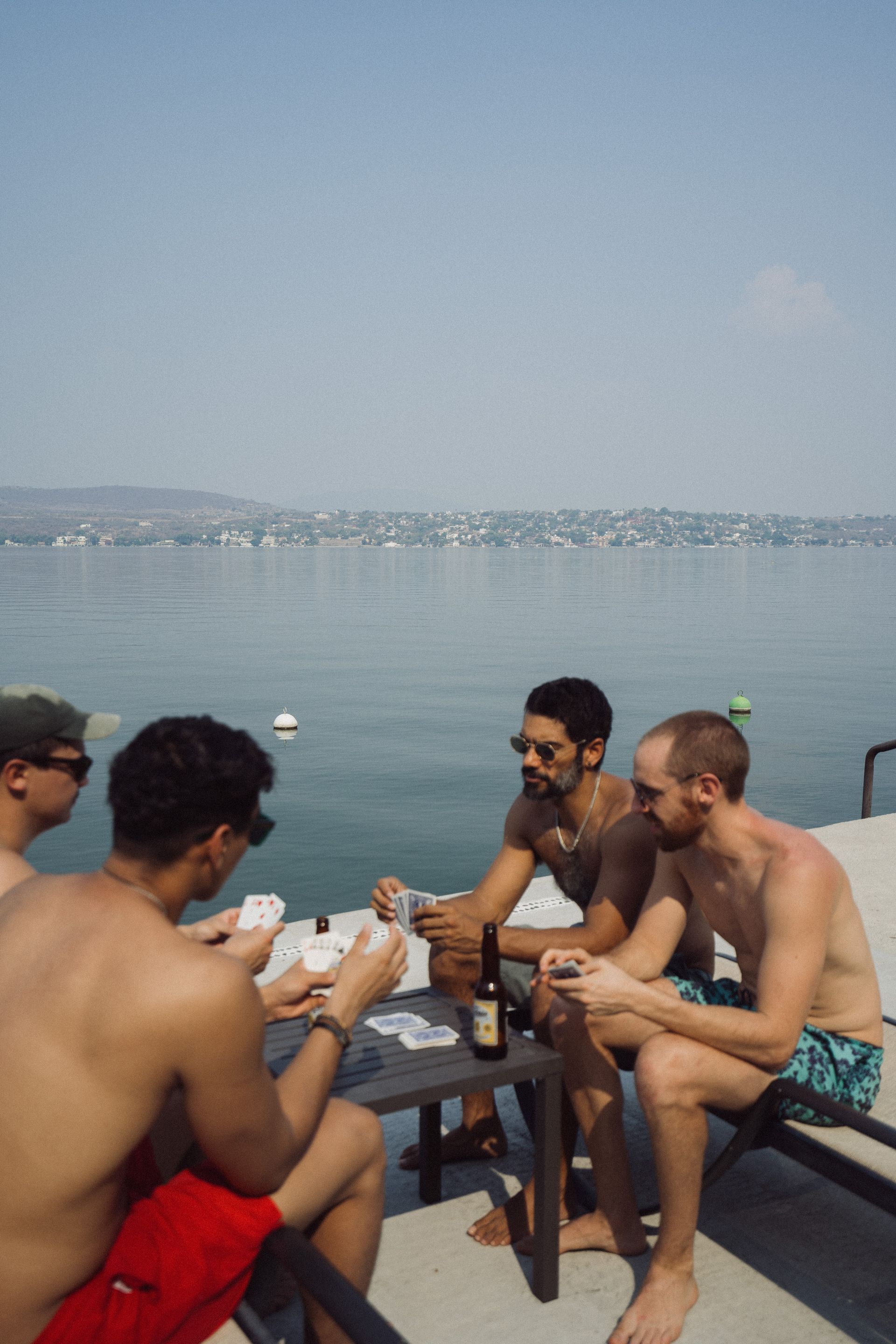 Cinco hombres sin camisa juegan a las cartas al aire libre junto al agua en un día soleado.