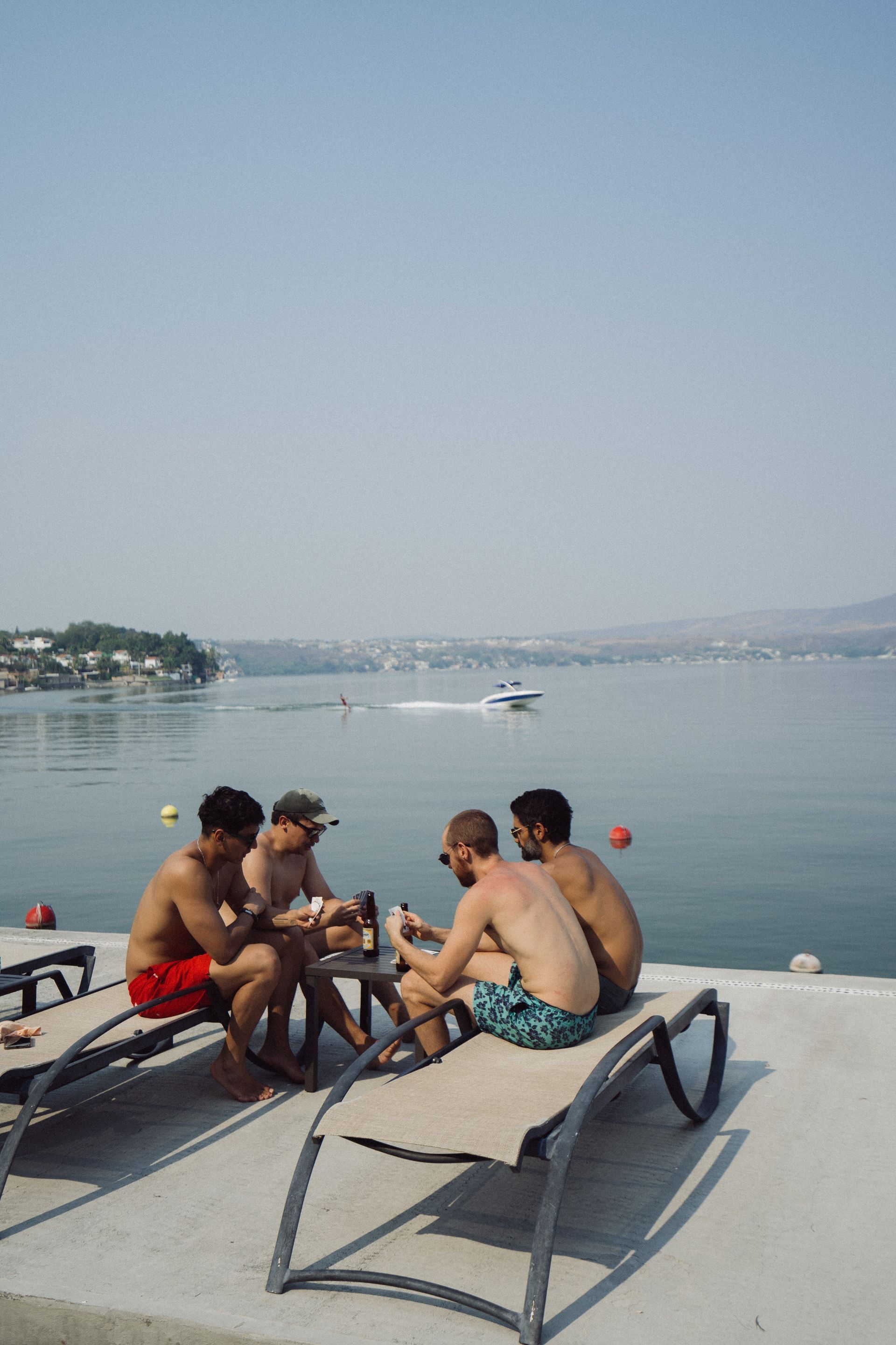 Cuatro hombres sin camisa están sentados en sillas reclinables, bebiendo junto a un lago en un día soleado.