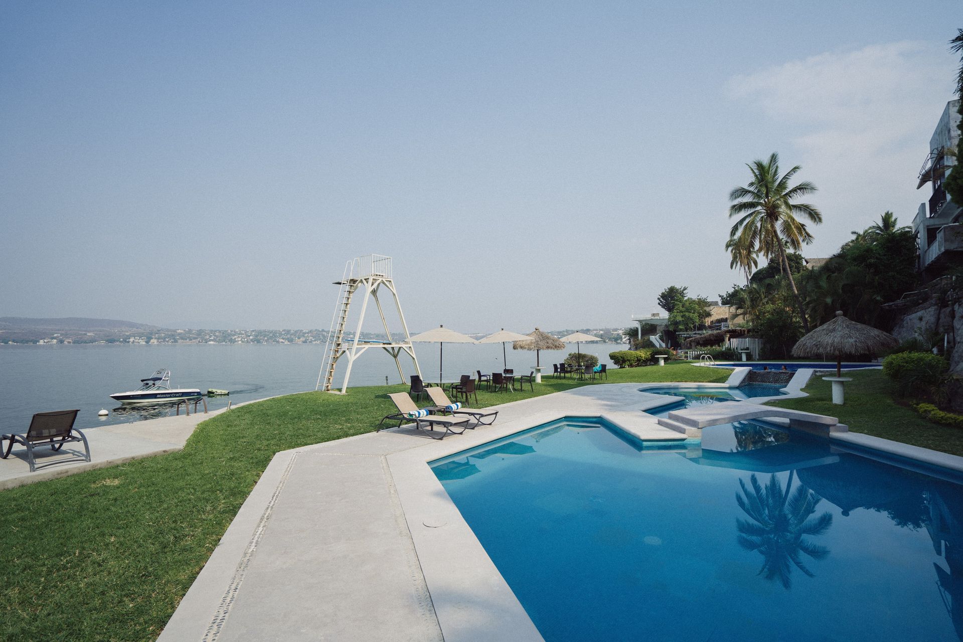 Vista a la piscina con agua turquesa, palmeras y un lago a lo lejos. Día soleado.