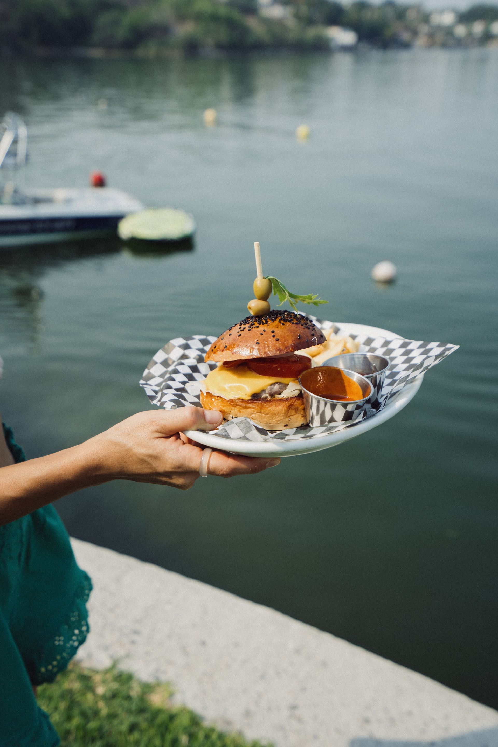 Persona sosteniendo un plato con una hamburguesa gourmet y papas fritas cerca de un cuerpo de agua.