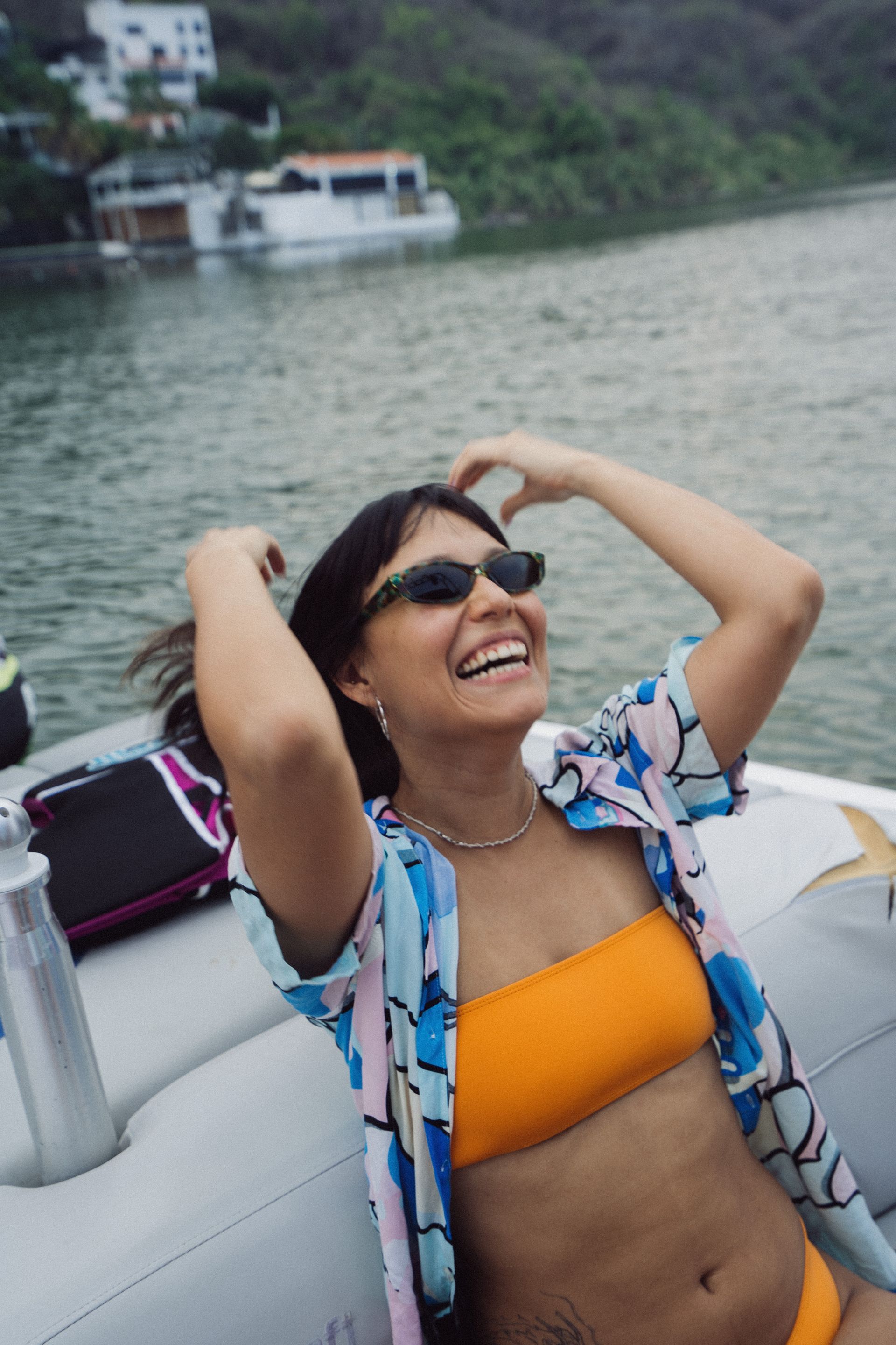 Una mujer en un barco sonríe, con una camiseta naranja, una camisa estampada y gafas de sol.