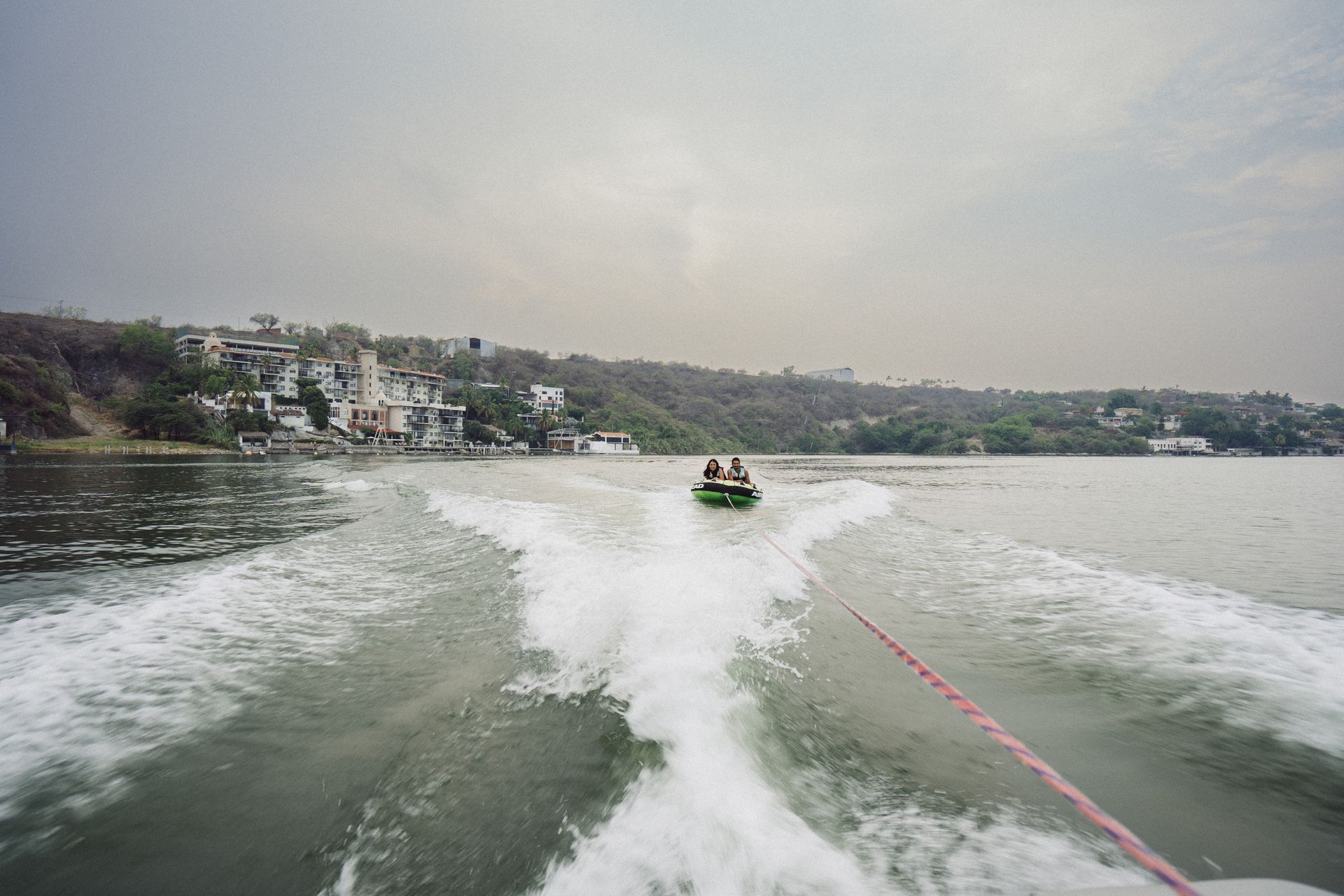 Una persona en un tubo arrastrado por un bote en un lago, con casas en la orilla lejana bajo un cielo nublado.