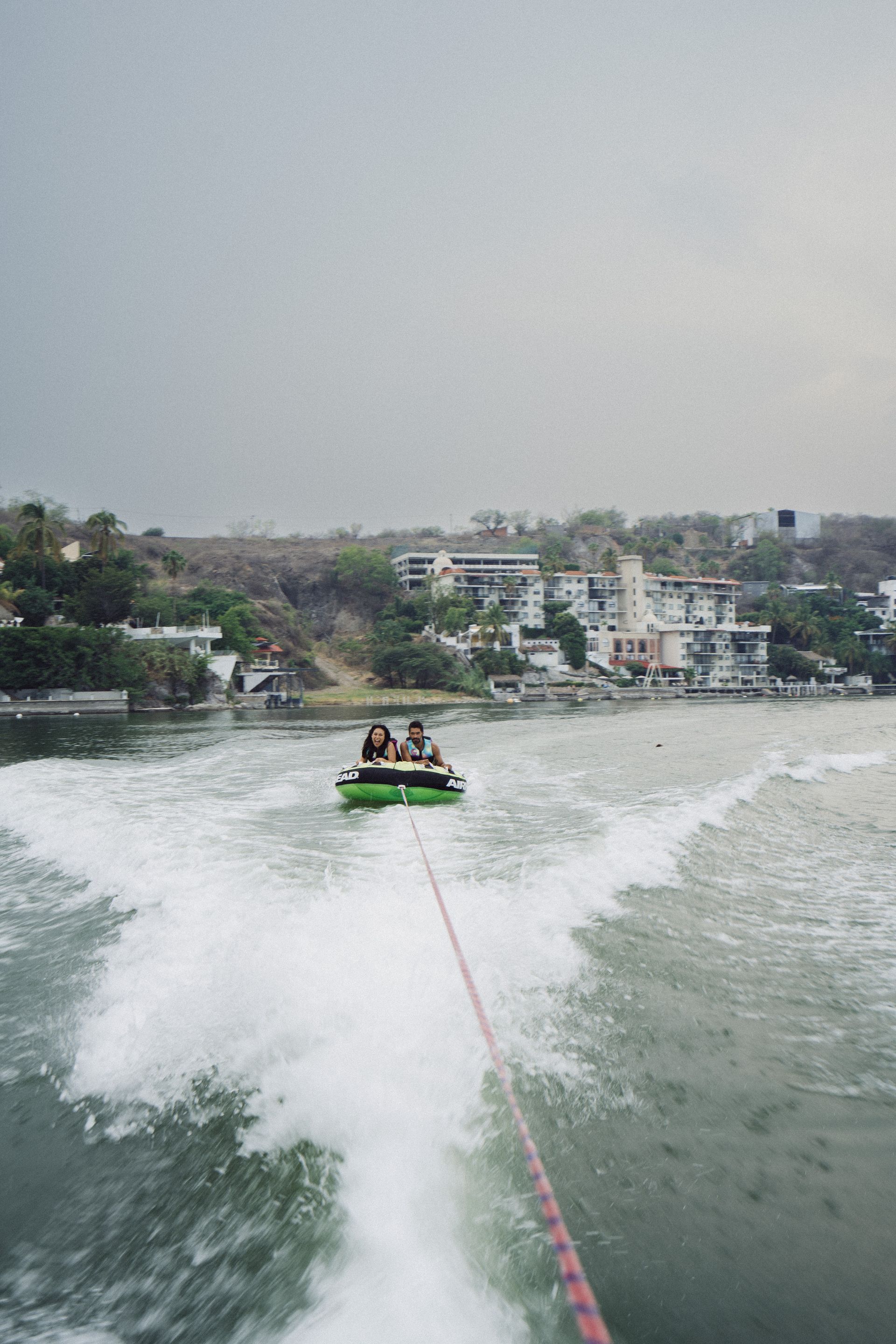 Un barco remolca a tres personas en un tubo inflable verde, en el agua, cerca de edificios bajo un cielo nublado.