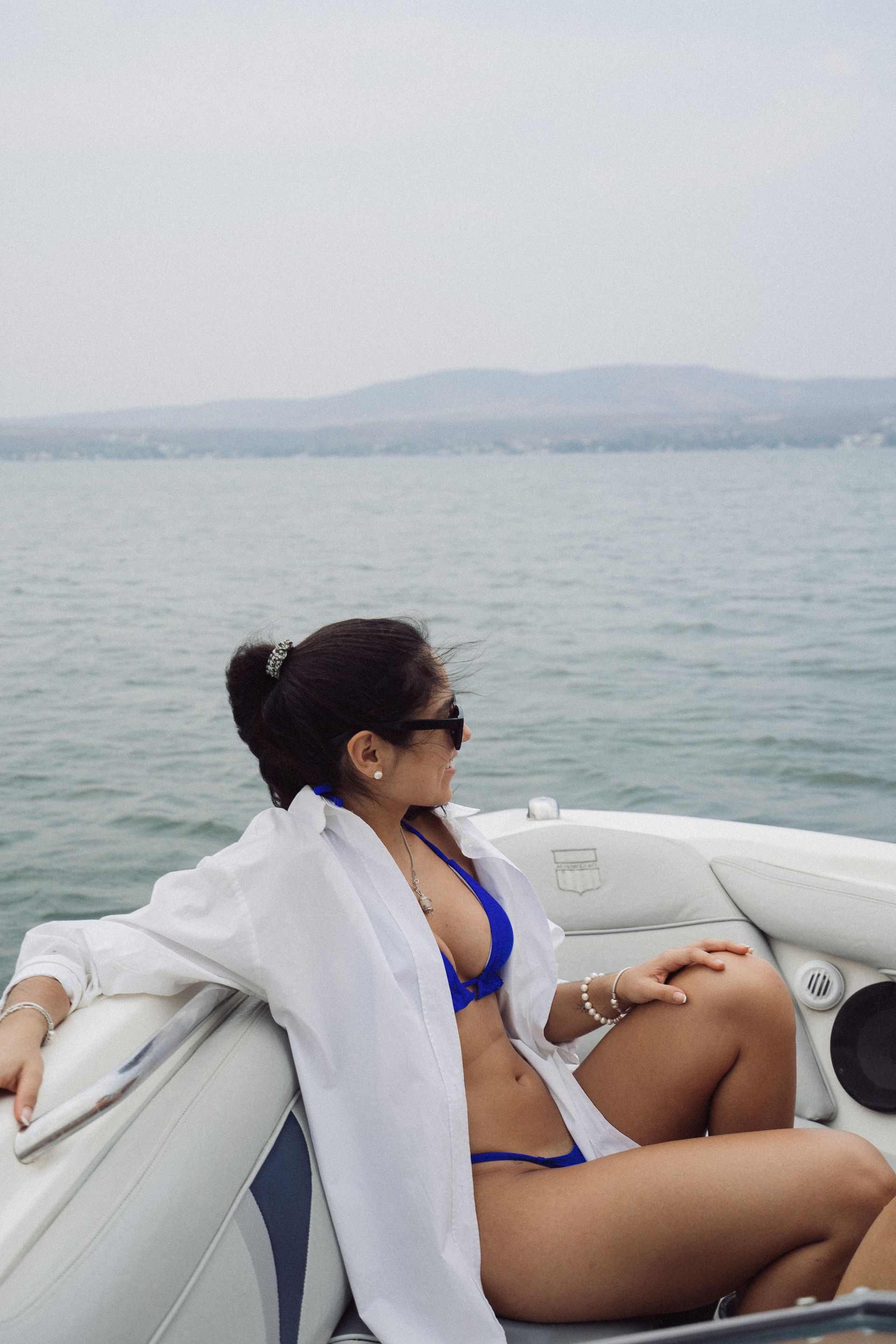 Mujer en bikini azul y camisa blanca en un barco, mirando hacia el agua.