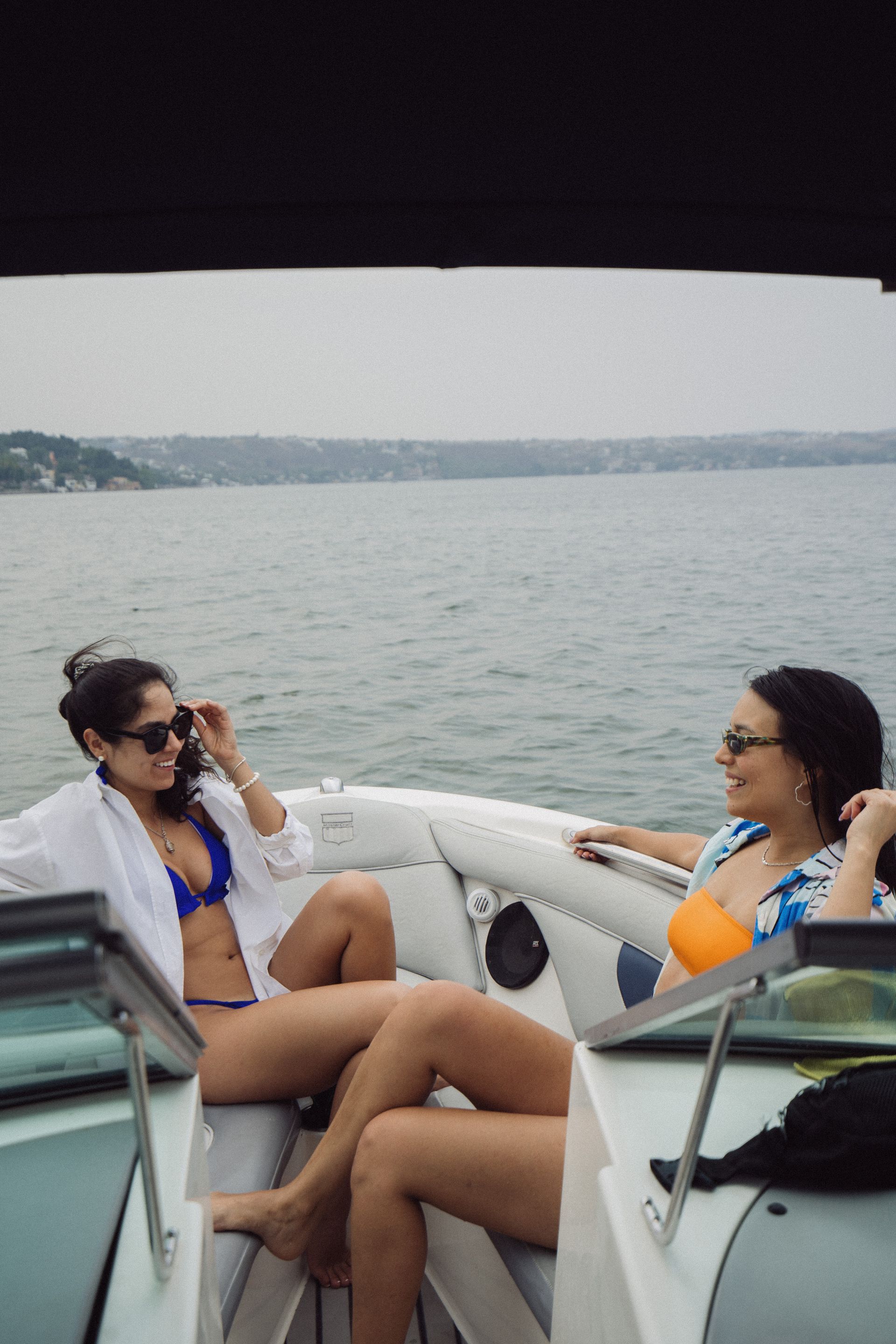 Dos mujeres se relajan en un bote en un lago, una con bikini azul y la otra con un top estampado. Cielo nublado.