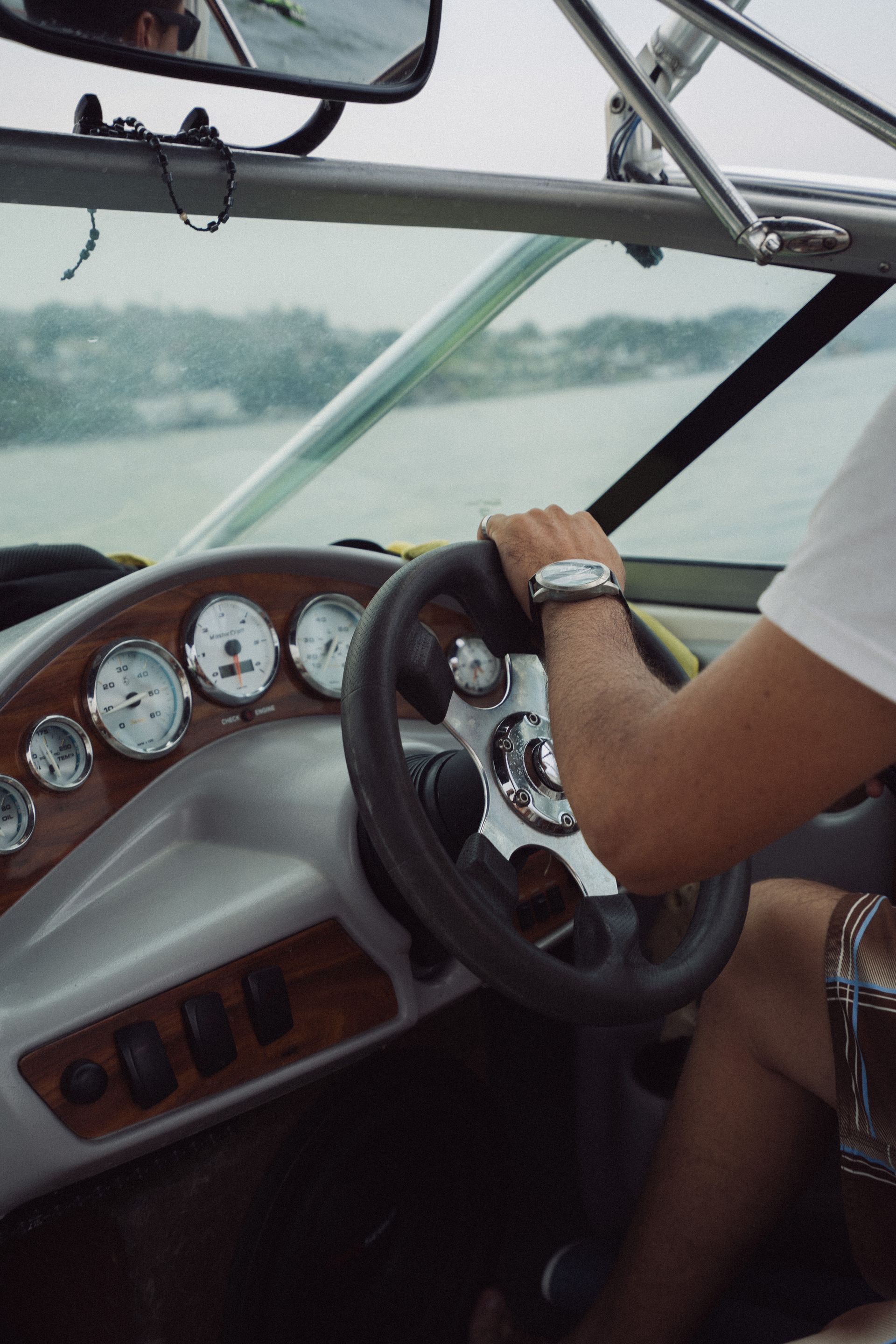 Una persona dirige un barco con la mano en el timón. Tablero con indicadores y un lago al fondo.