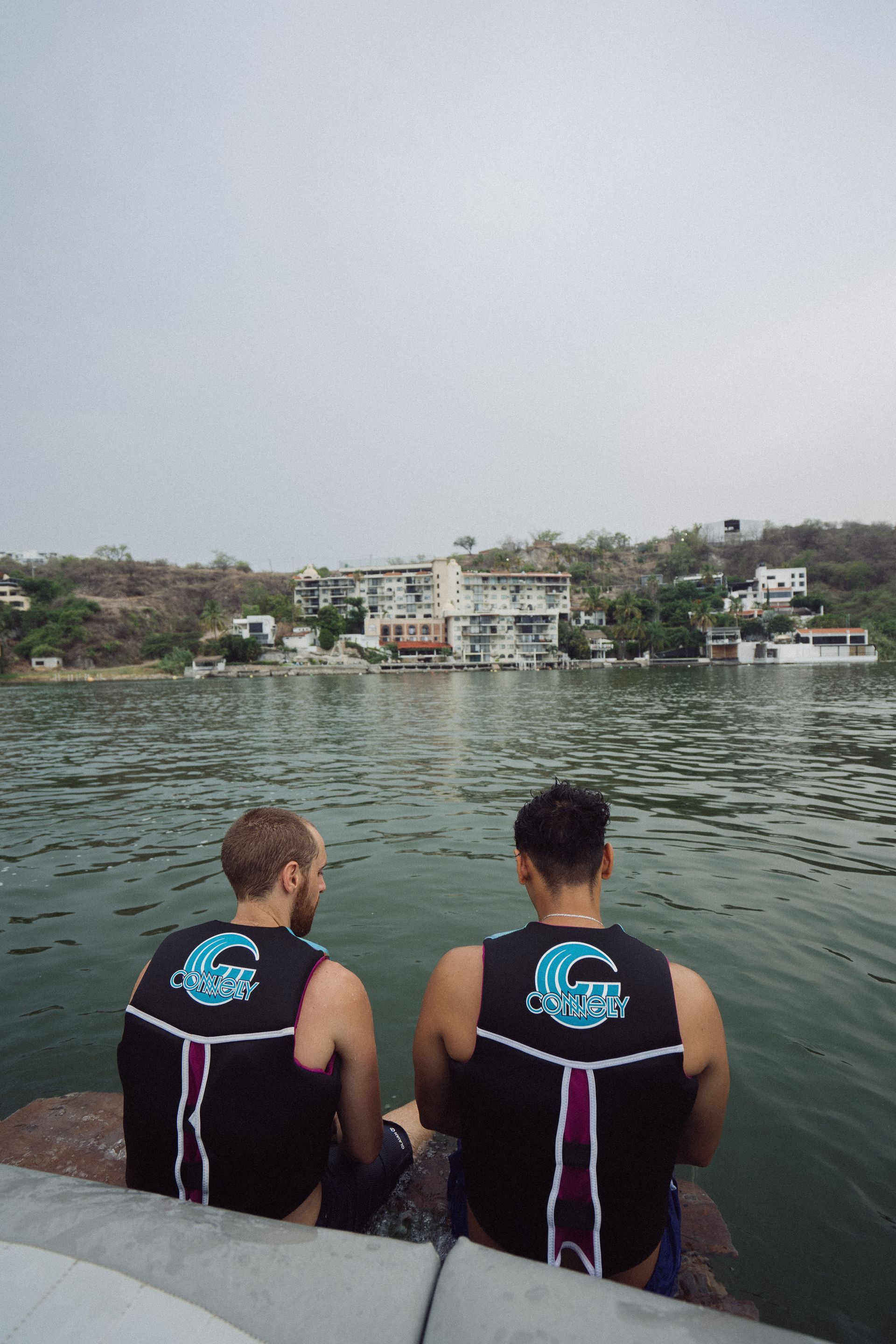 Dos hombres con chalecos salvavidas están sentados en un bote, mirando un pueblo junto al lago bajo un cielo nublado.