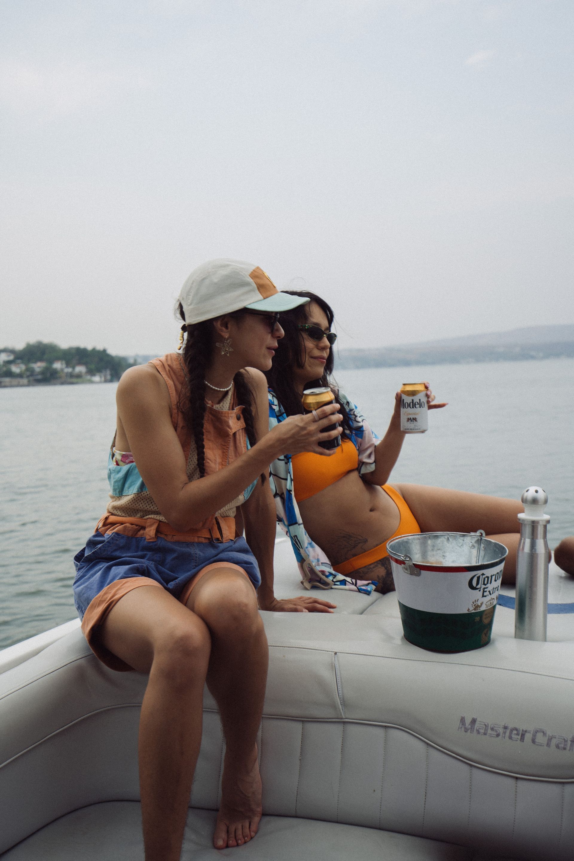 Dos mujeres en un barco, bebiendo de latas. Una lleva sombrero, la otra gafas de sol. Cielo nublado.