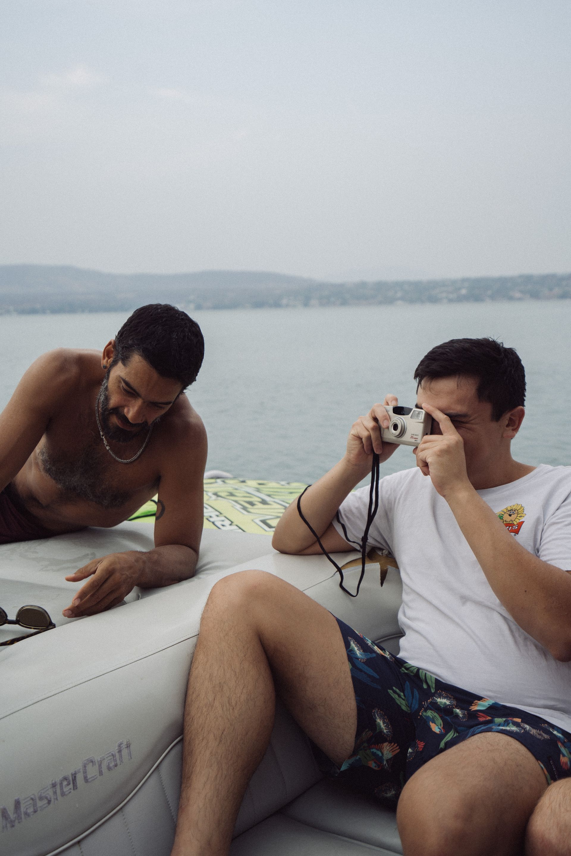 Dos hombres en una balsa inflable, uno toma fotos con la cámara, el otro descansa, el agua y la orilla lejana al fondo.
