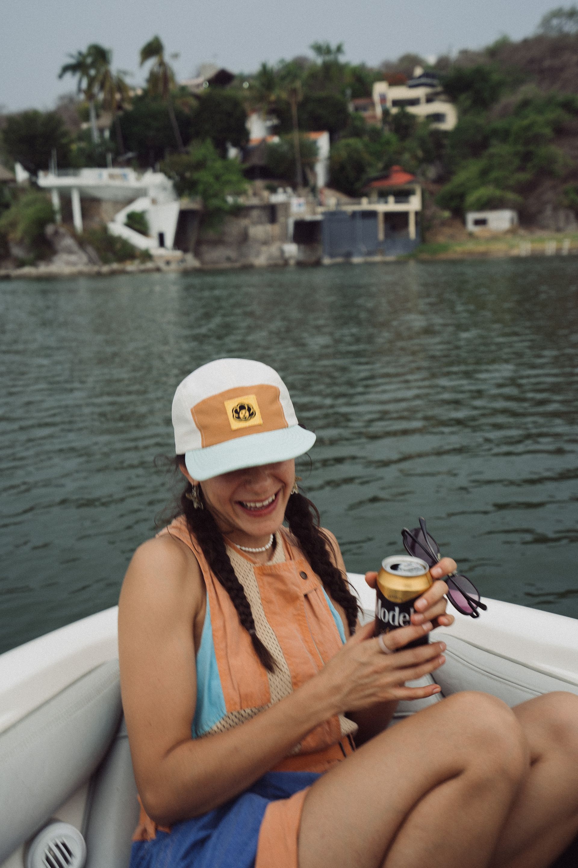 Mujer en un barco sonriendo, sosteniendo una bebida, con casas en una ladera al fondo.