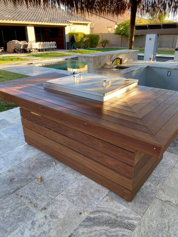 Wooden outdoor grill island with a light-colored countertop. The island is near a pool and patio.