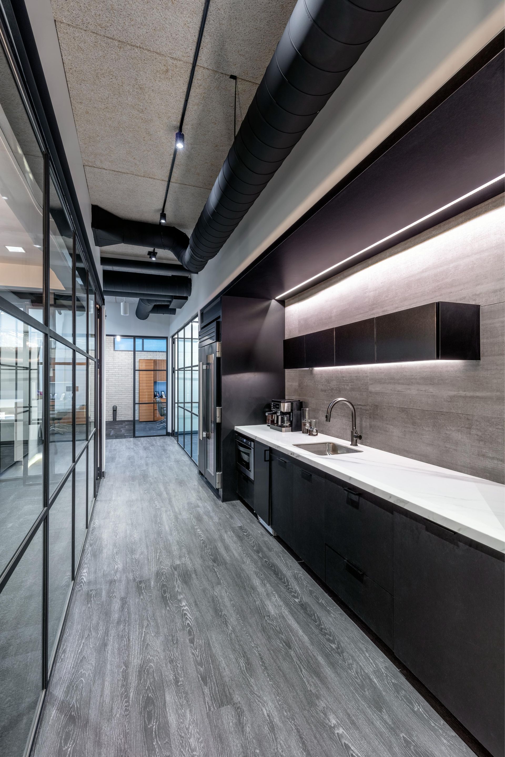 Office hallway with kitchen area. Dark cabinetry, white countertop, gray flooring. Glass wall to the left.
