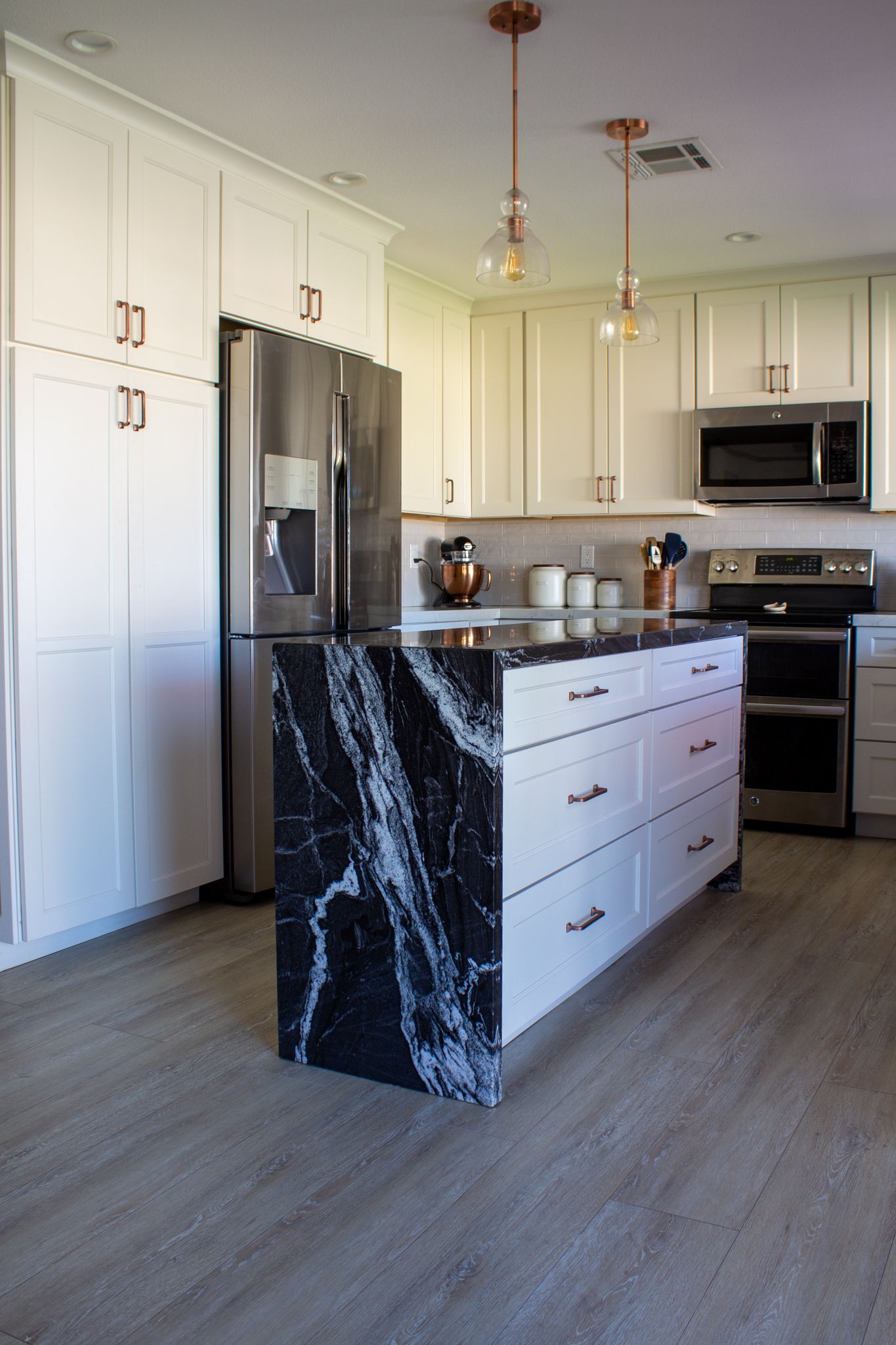 Kitchen with white cabinets, stainless steel appliances, black marble island, and light wood-look flooring.