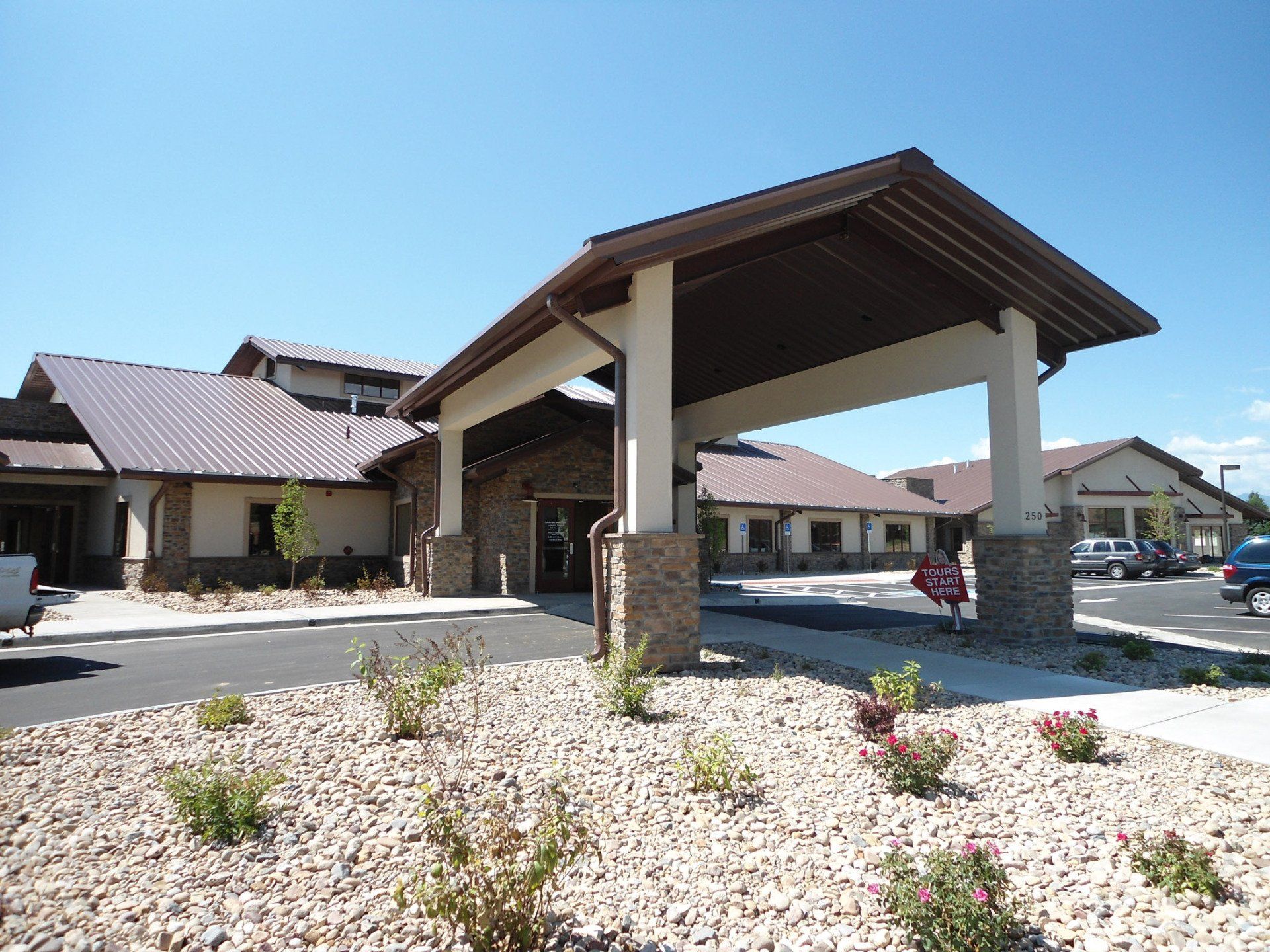 Brown-roofed building with covered entrance, beige walls, and landscaping under a blue sky.