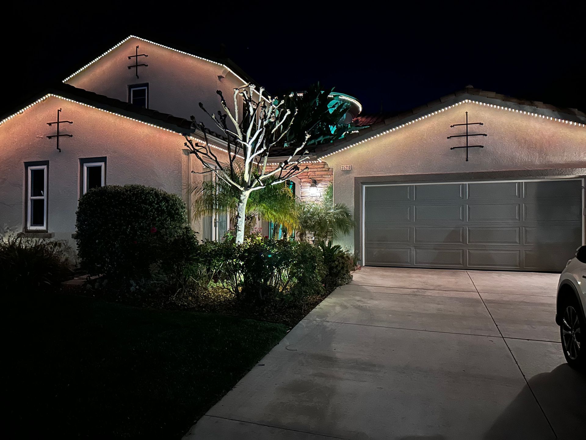House at night, decorated with white Christmas lights. Garage and front door illuminated; dark driveway.