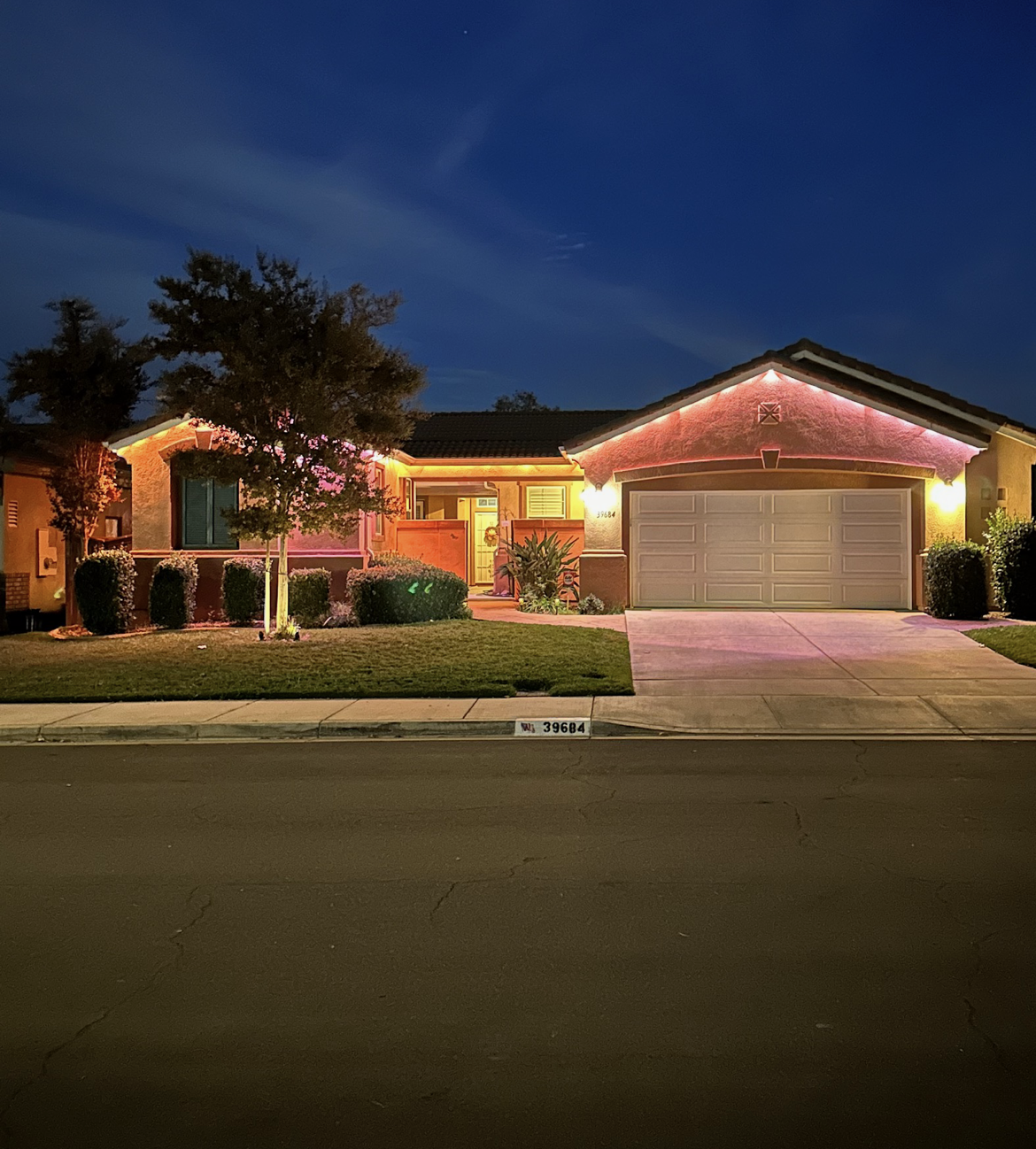 A house lit up with pink lights along the roofline and on a tree in the yard at dusk.