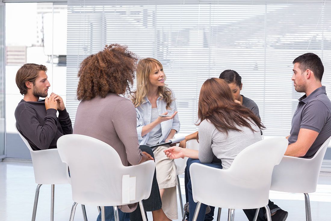 Small group in a bright office meeting, seated in a circle and talking with notes in hand.
