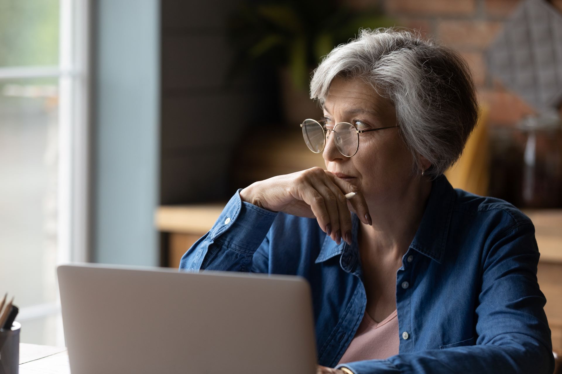 Person in a blue shirt sitting at a laptop, looking thoughtfully out a window.