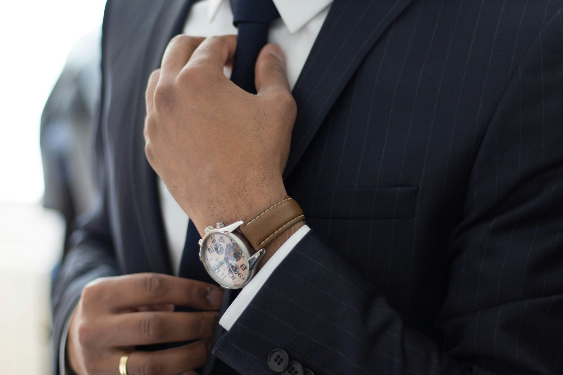 Man in a navy suit adjusting his tie, wearing a wristwatch and bracelets.