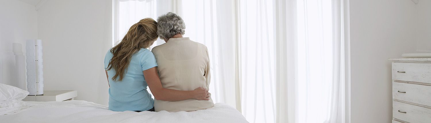 Two people sit on a bed, embracing and looking out a bright window with white curtains.