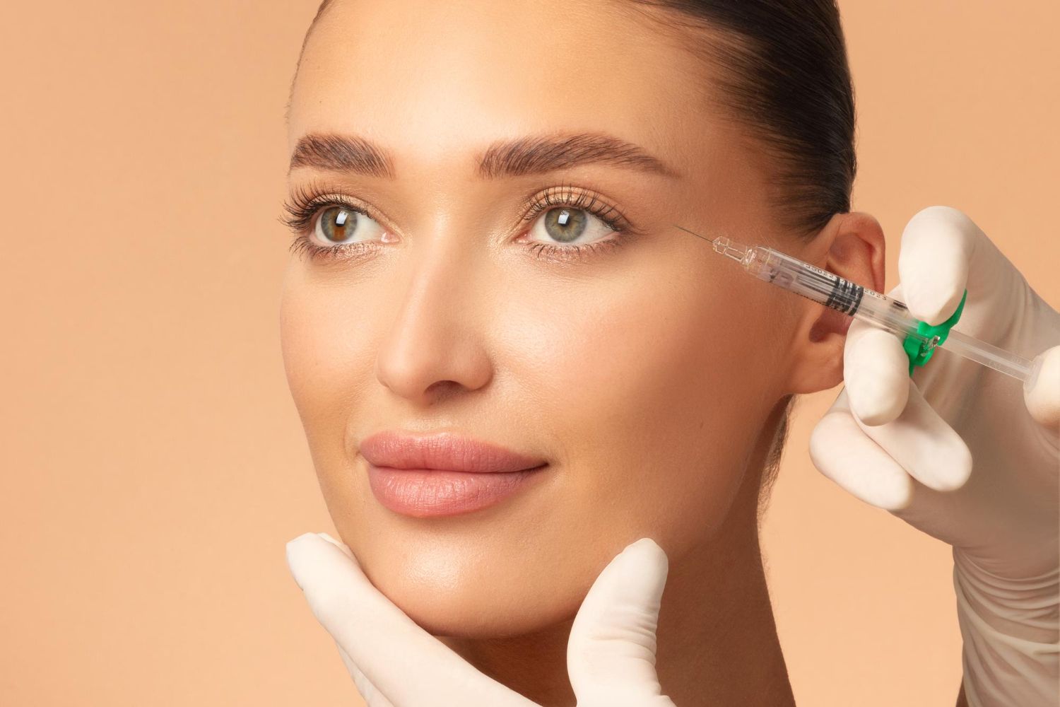 Woman receiving a facial injection near the eye; light skin, beige background, gloved hand.
