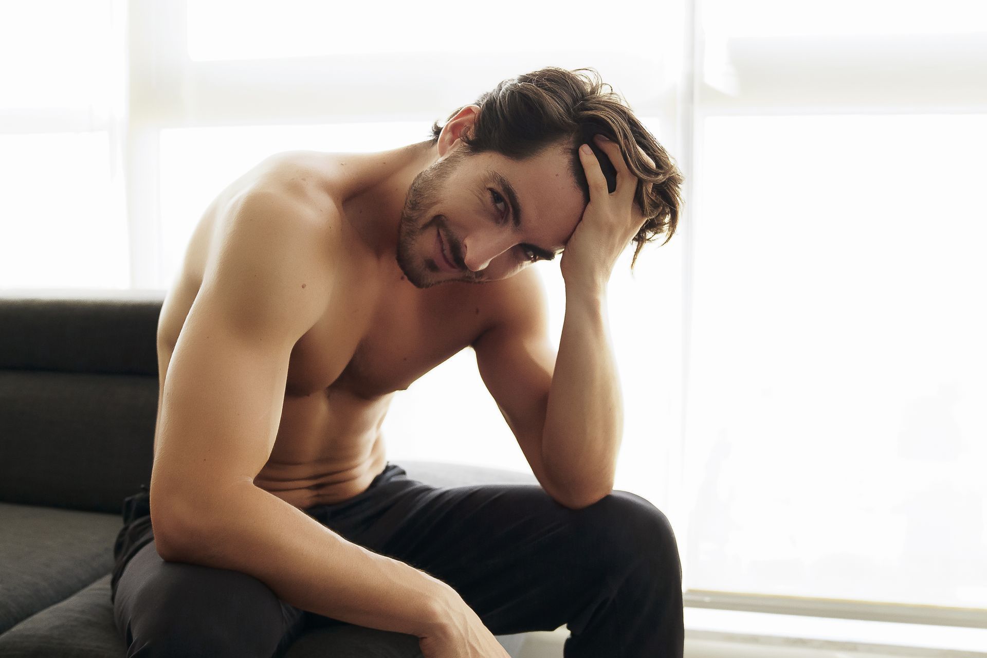 A shirtless man is sitting on a couch with his hand on his head.