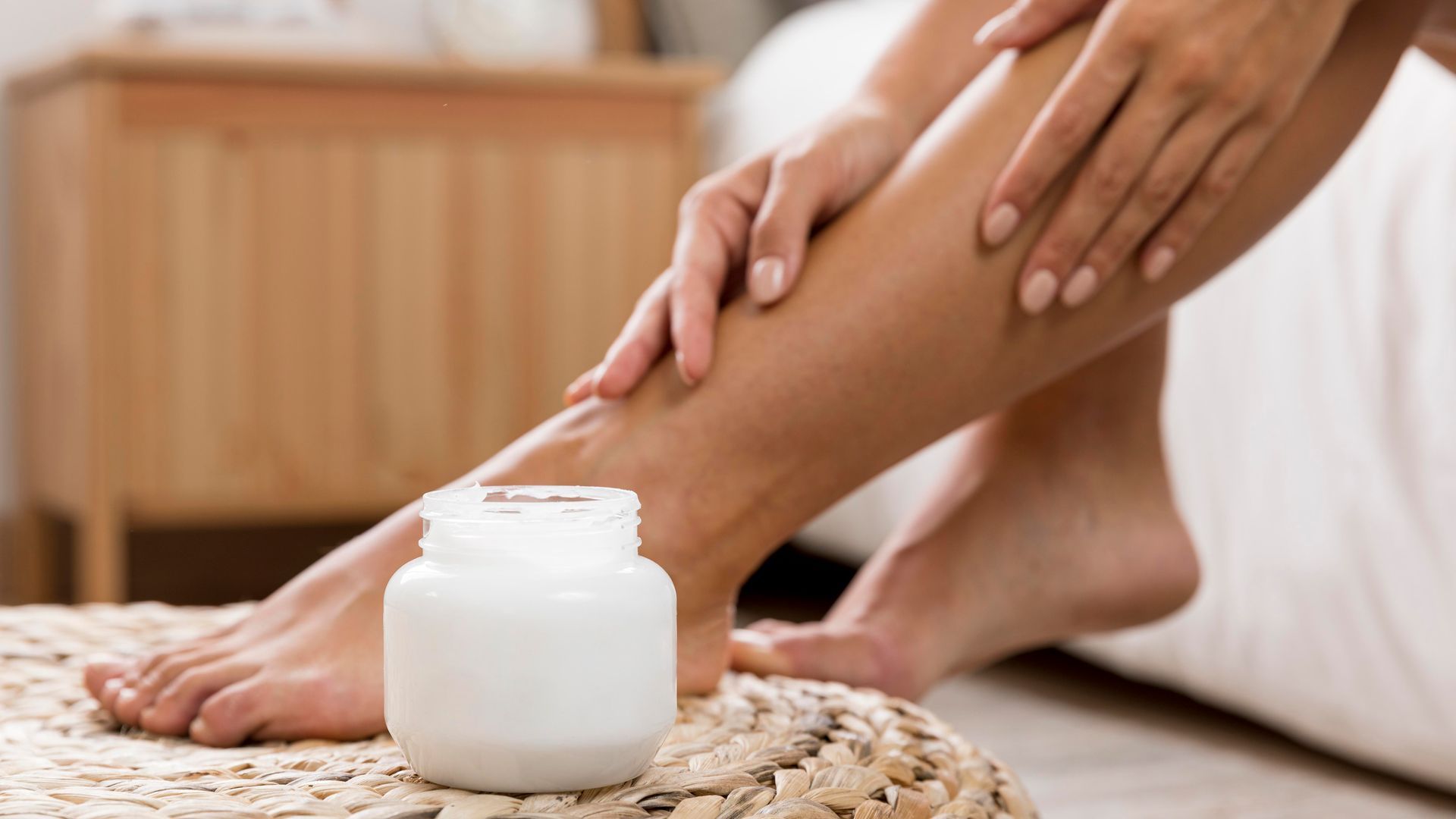 A woman is applying lotion to her legs next to a jar of lotion.