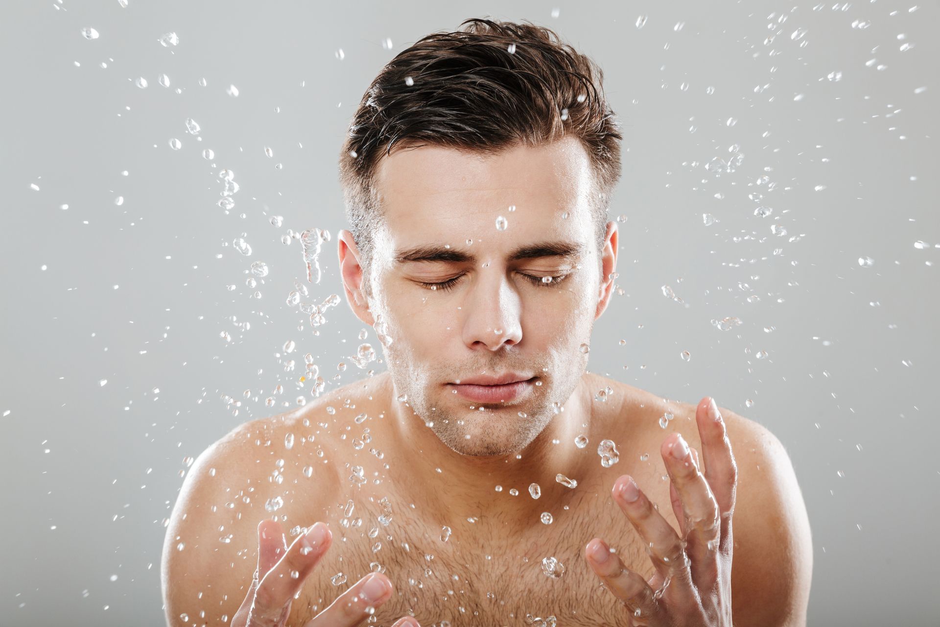 A shirtless man is washing his face with water.