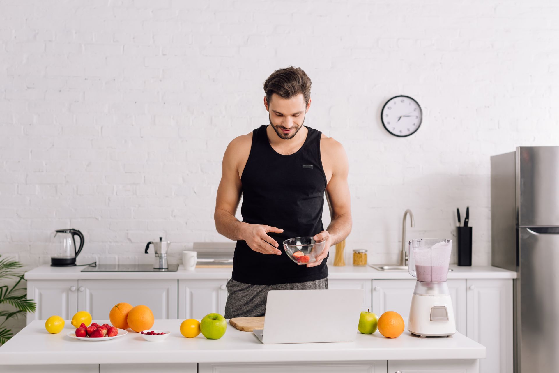 A man is standing in a kitchen preparing food in front of a laptop.