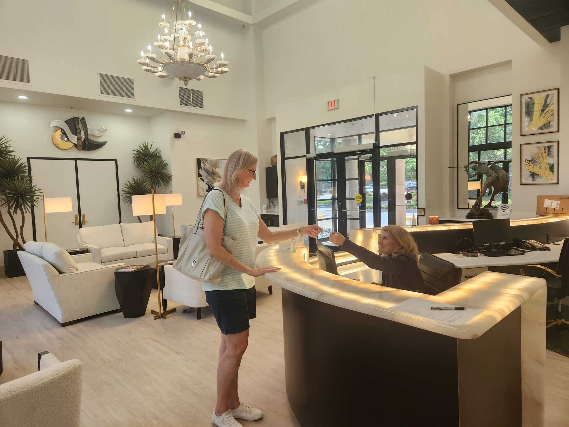 Woman at reception desk, handing document. Bright lobby with seating, chandelier, and a second woman behind the desk.