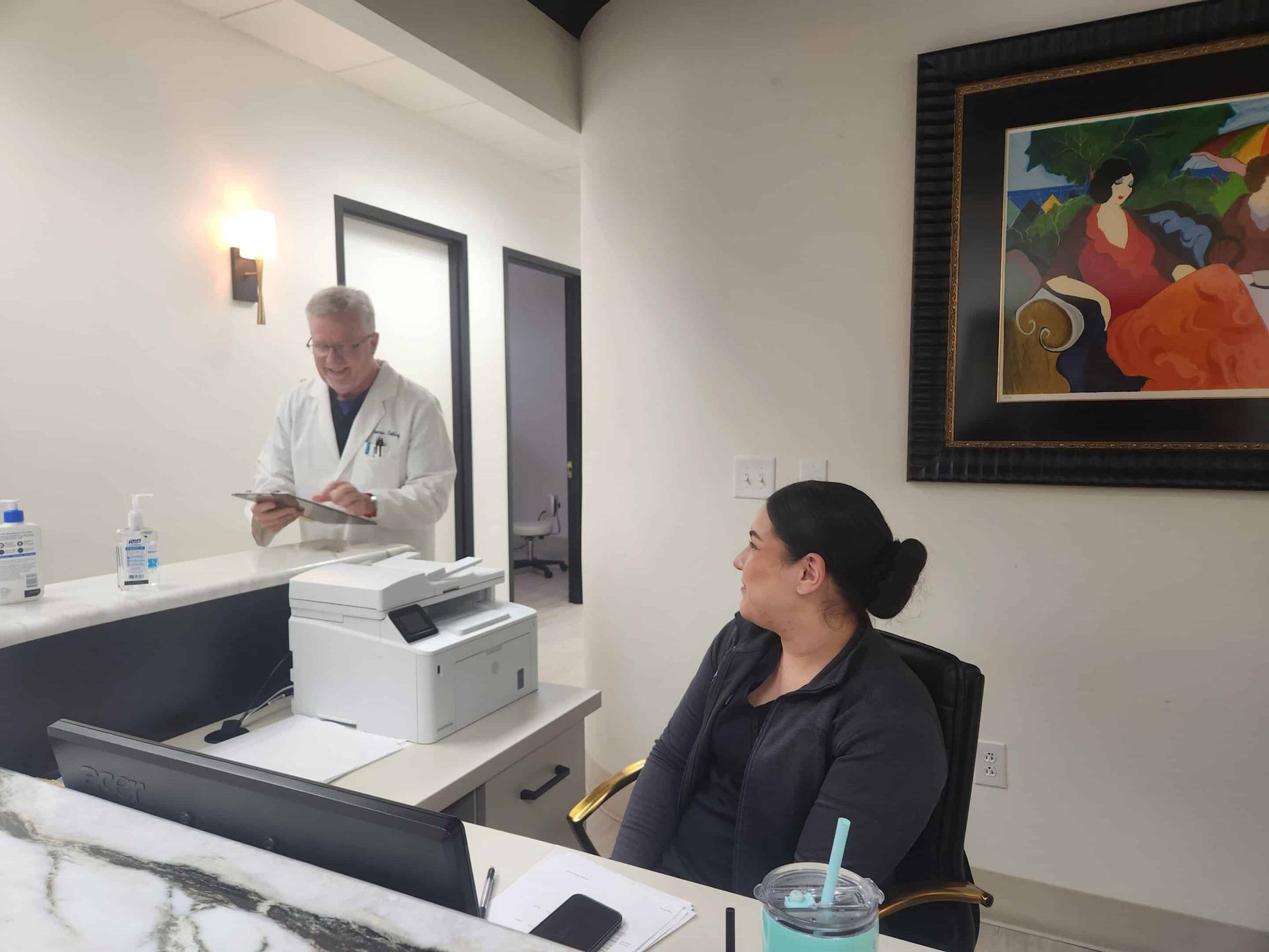 Doctor in white coat at a reception desk, speaking to seated staff member. Office setting, artwork on wall.