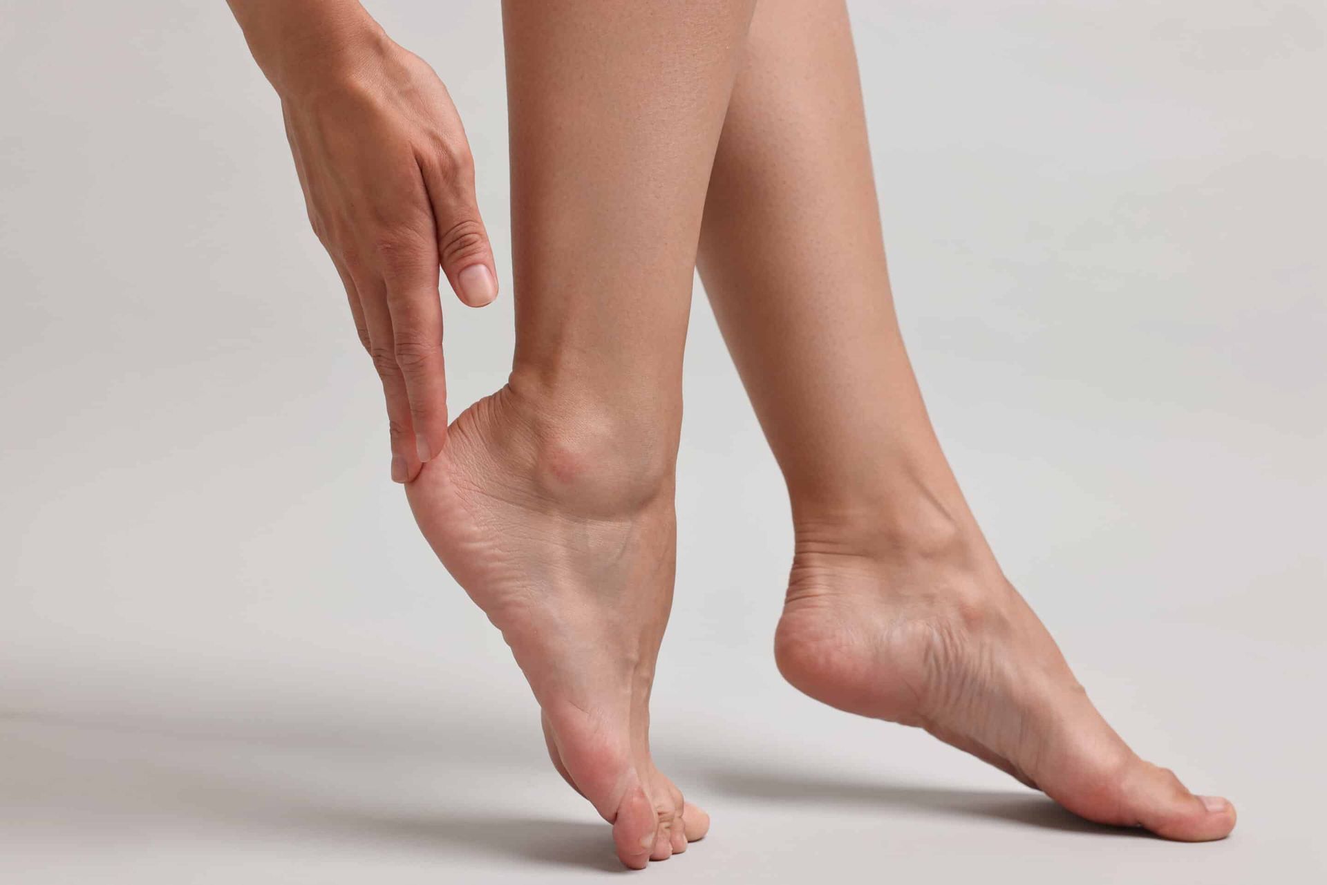 Person's bare feet, one foot raised, hand touching the heel, on a neutral gray background.