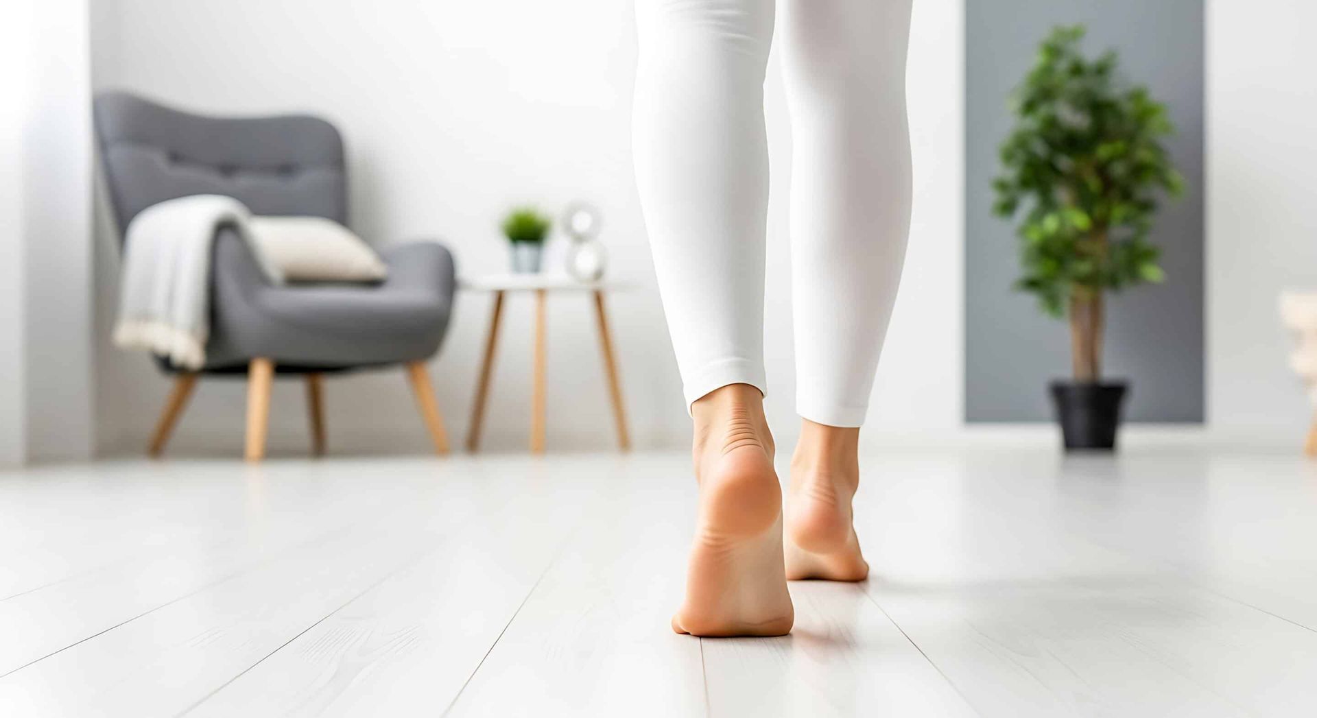 Person walking barefoot on a white floor toward a living area with a chair, table, and plant.