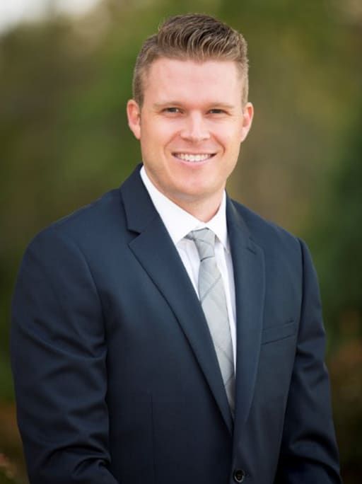 Man in navy suit, white shirt, and silver tie smiles against a blurred green background.